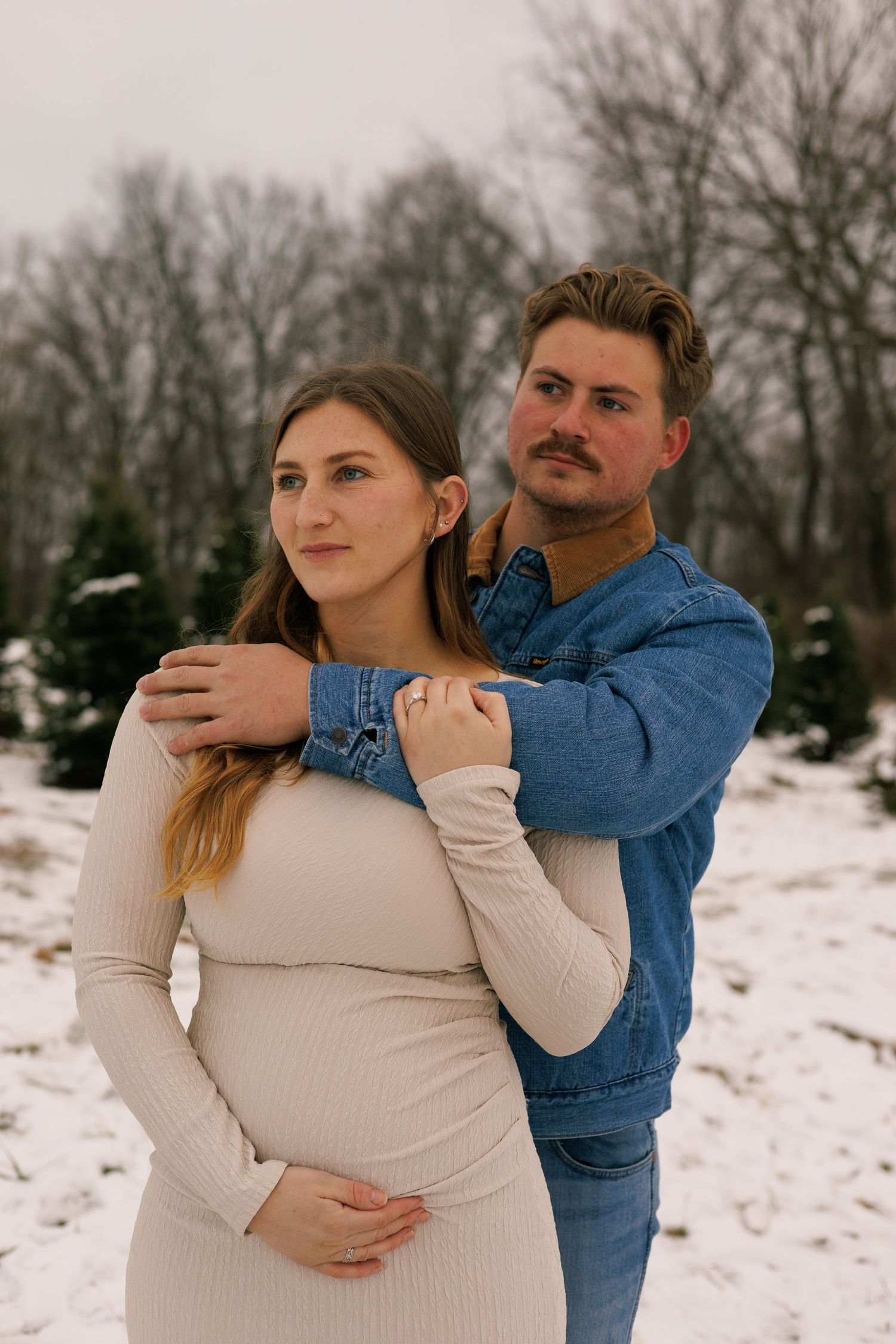 A couple embraces in a snowy winter setting wearing a beige sweater and denim jacket in Northeast Ohio.