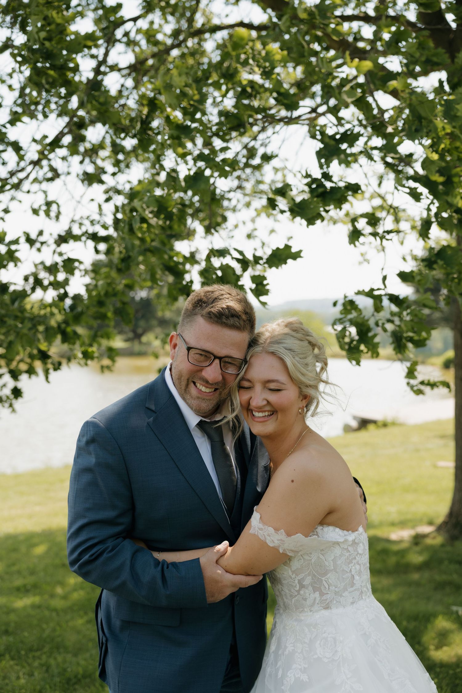 Laughing bride and groom embrace outdoors under green tree by lake, groom in navy suit with glasses.