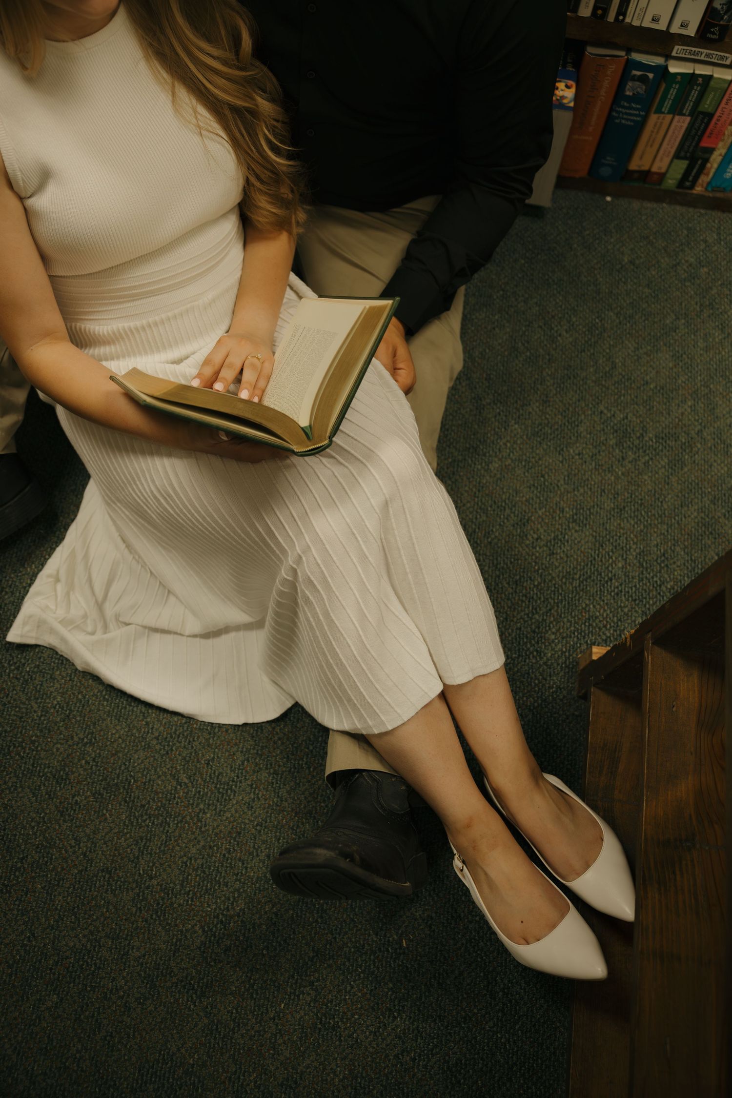 Couple sitting together on steps in a library, woman in white ribbed dress and flats holding an open book.