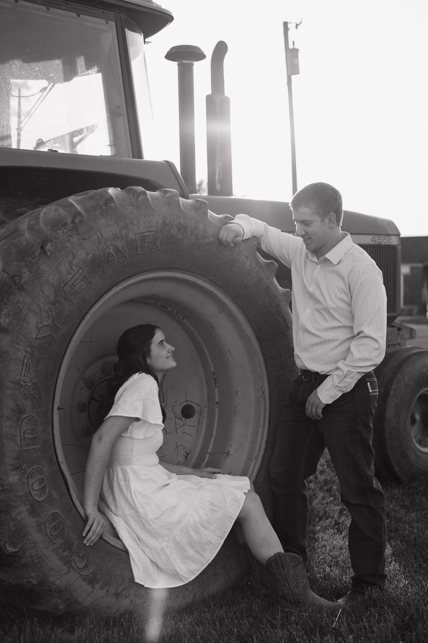 Black and white photo of a couple sharing a romantic moment beside a large tractor tire on a farm.