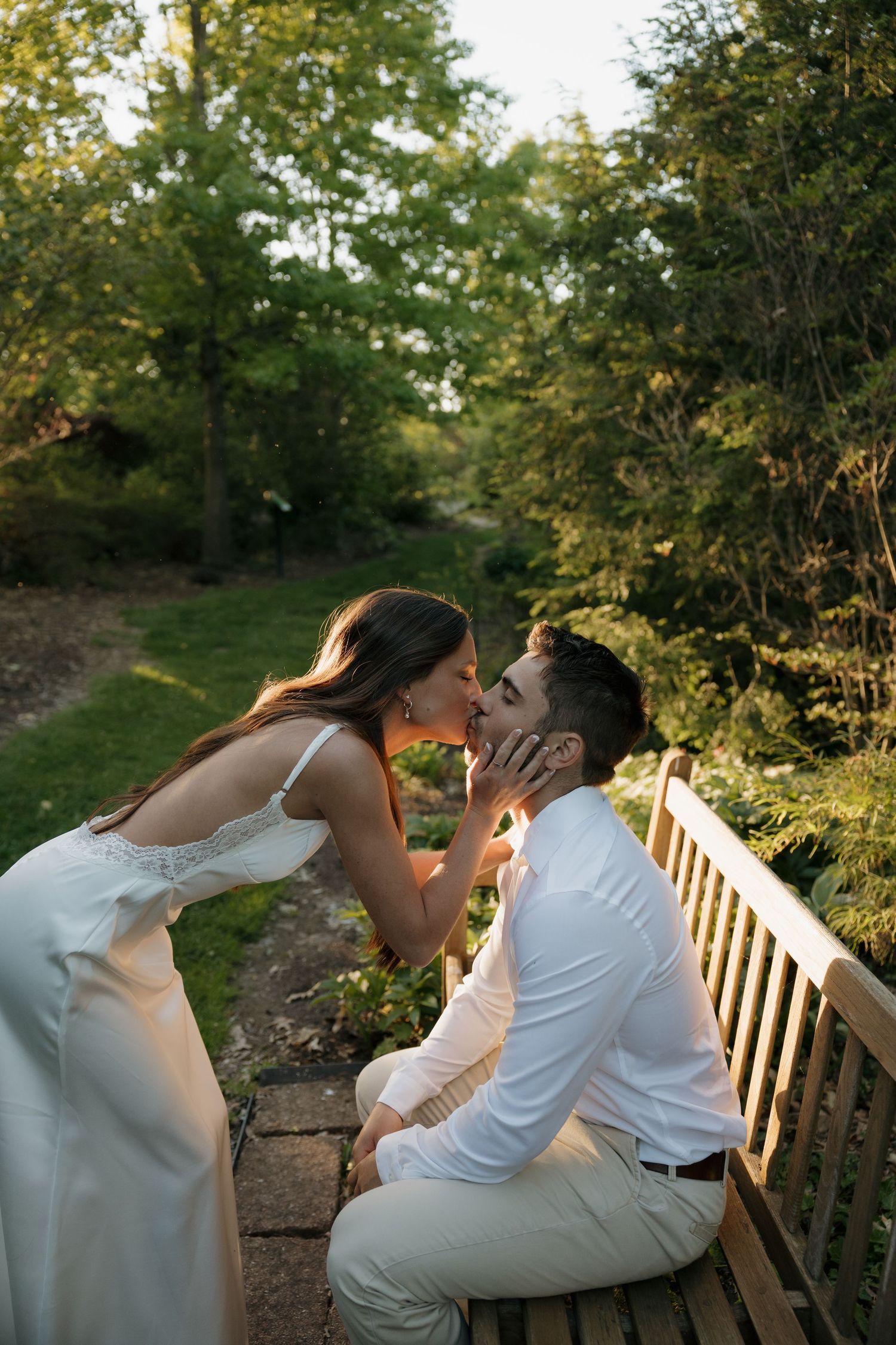 A bride in a white gown leans in to kiss her groom seated on a wooden bench in a lush garden setting.