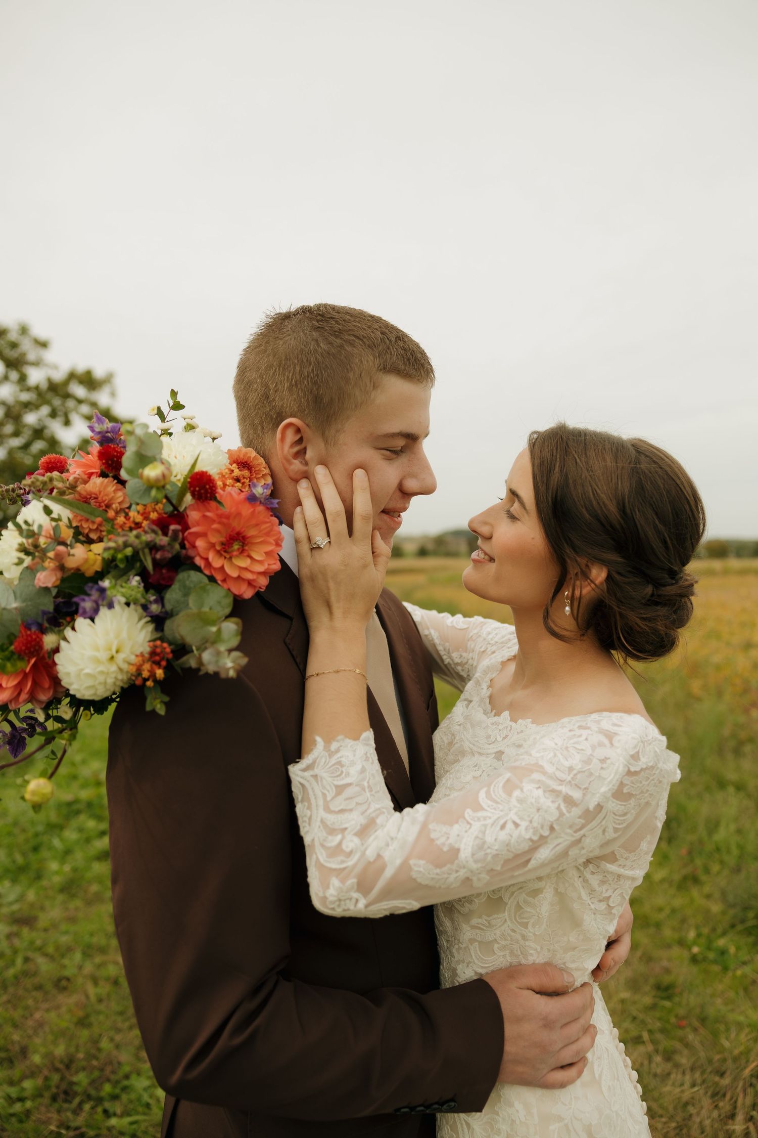 Bride in a lace gown gazes lovingly at her groom in a brown suit while holding a colorful dahlia bouquet in a grassy field.