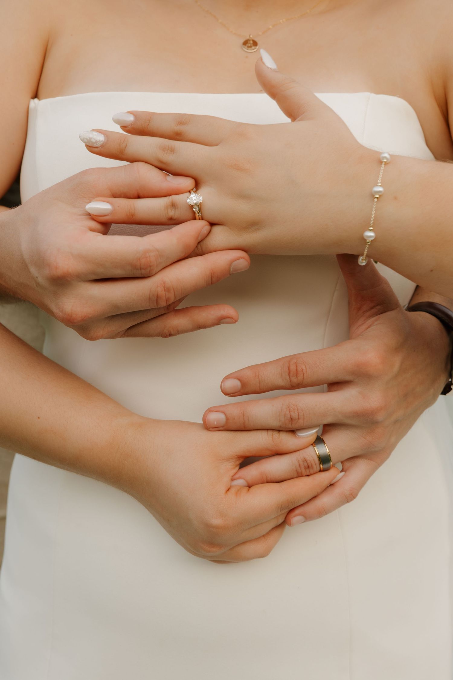 Close-up of bride and groom hands with wedding rings, pearl bracelet, white dress.