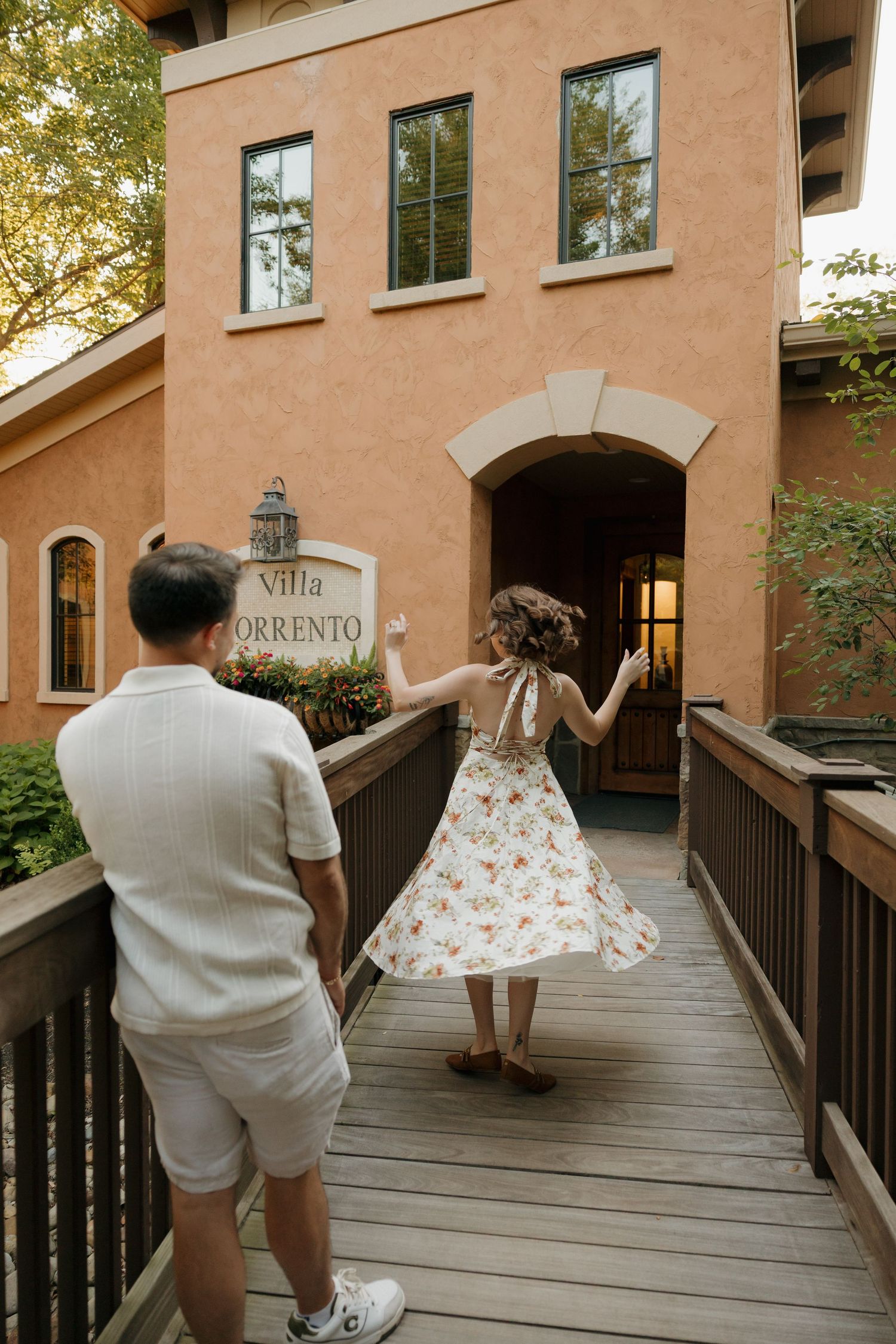 Couple walking toward Villa Sorrento entrance on wooden bridge, woman in floral dress twirling, man in white outfit behind her.