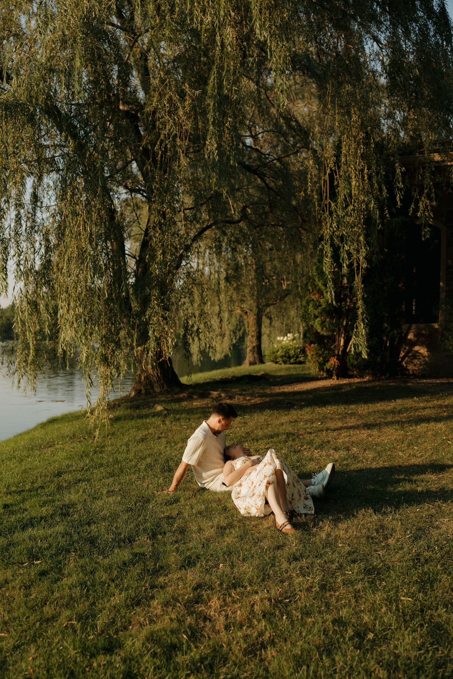 Couple relaxing on green grass under weeping willow trees by a serene lake in warm golden sunlight.
