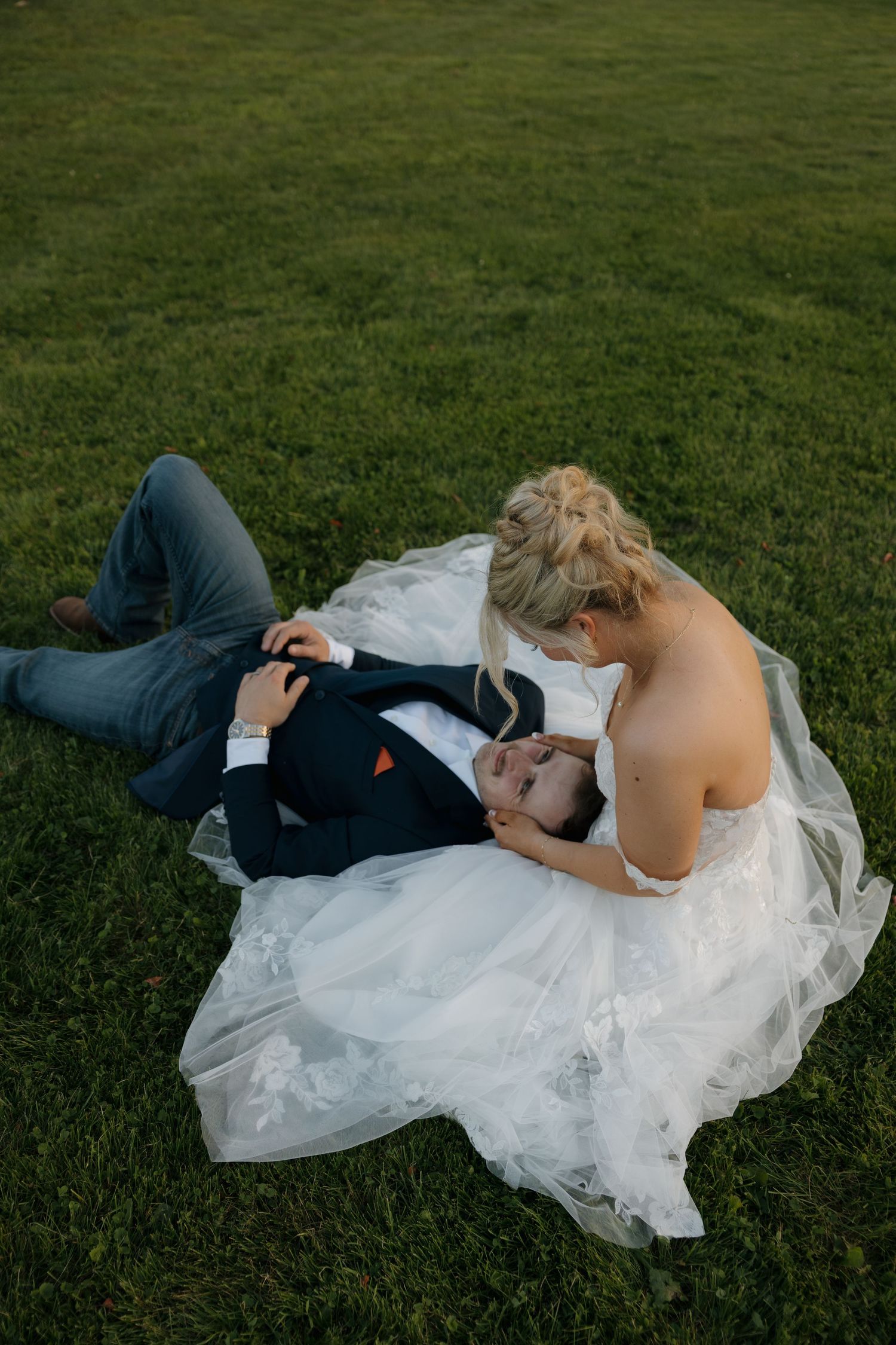 Bride in white gown sits beside groom lying on green grass in an intimate outdoor wedding photo.