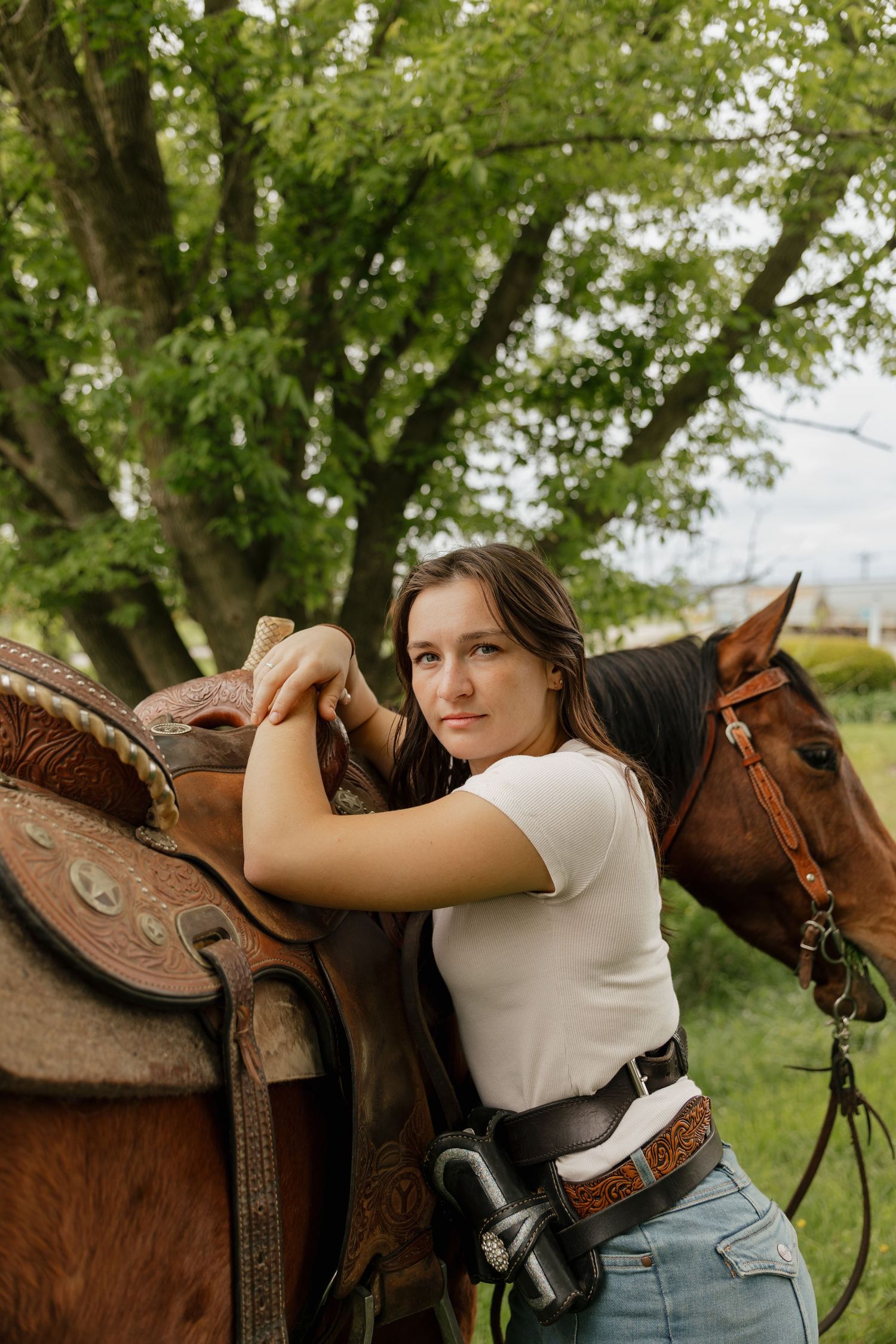 Young woman in white t-shirt and jeans leans on a Western saddle beside a chestnut horse under a green tree.