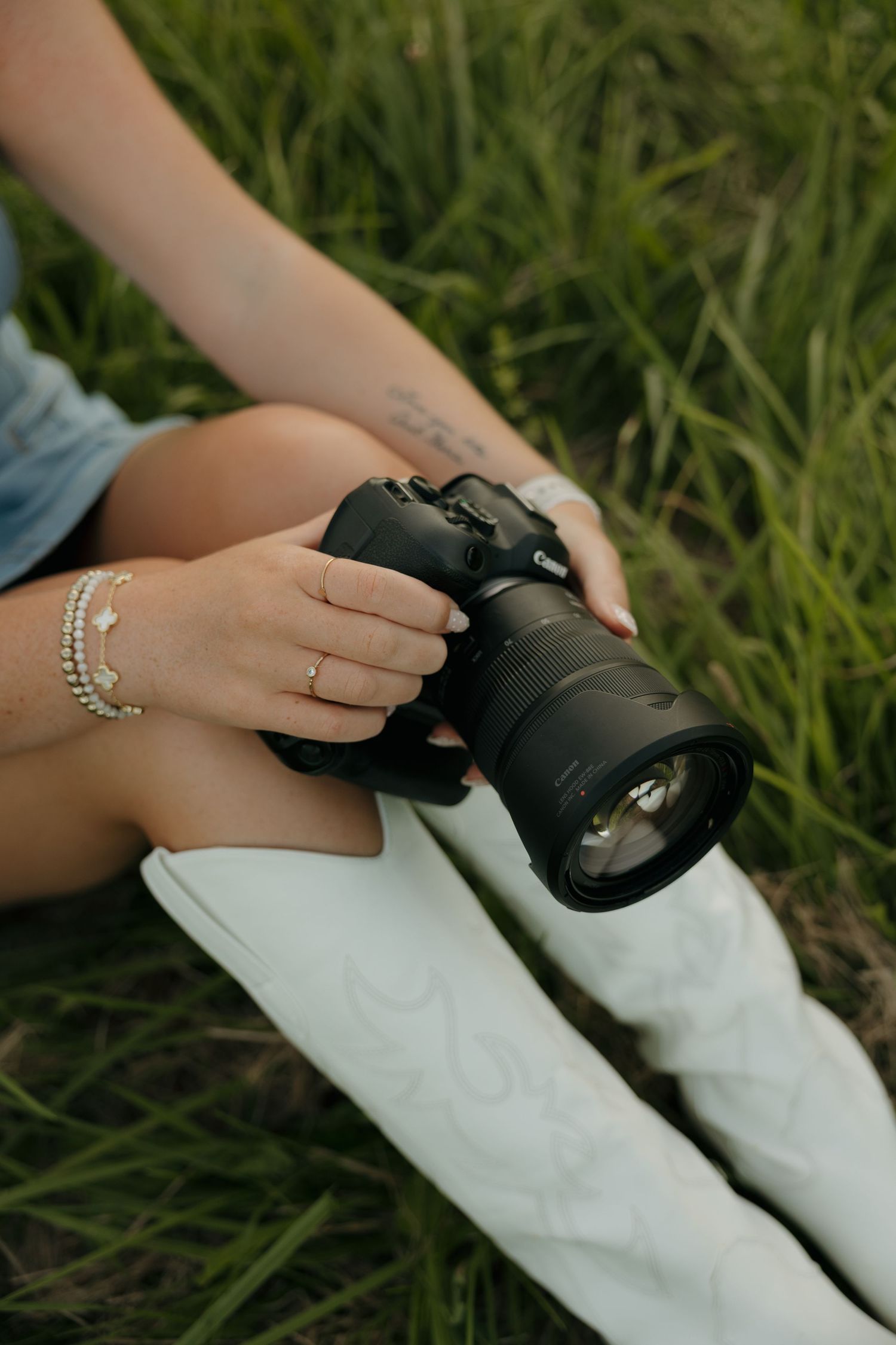 Woman sitting in green grass holding a Canon DSLR camera, wearing white cowboy boots, denim shorts, and silver bracelets.