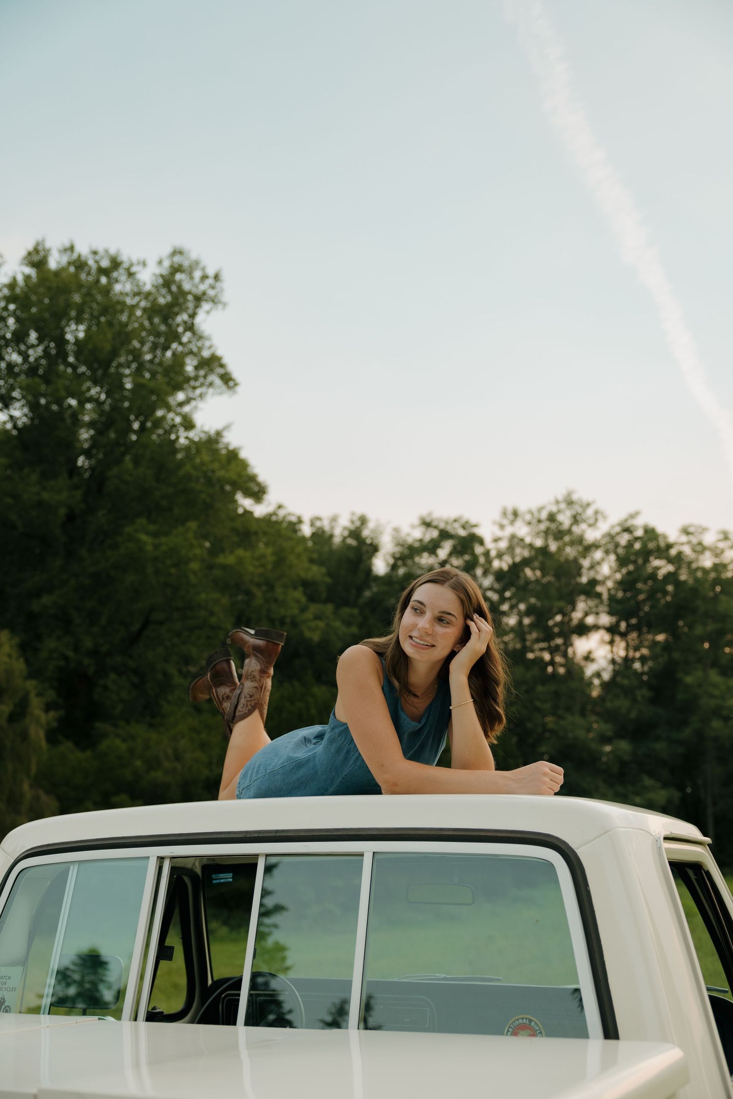 Young woman in denim overalls lounging on top of a white vintage van, smiling outdoors with trees in background.