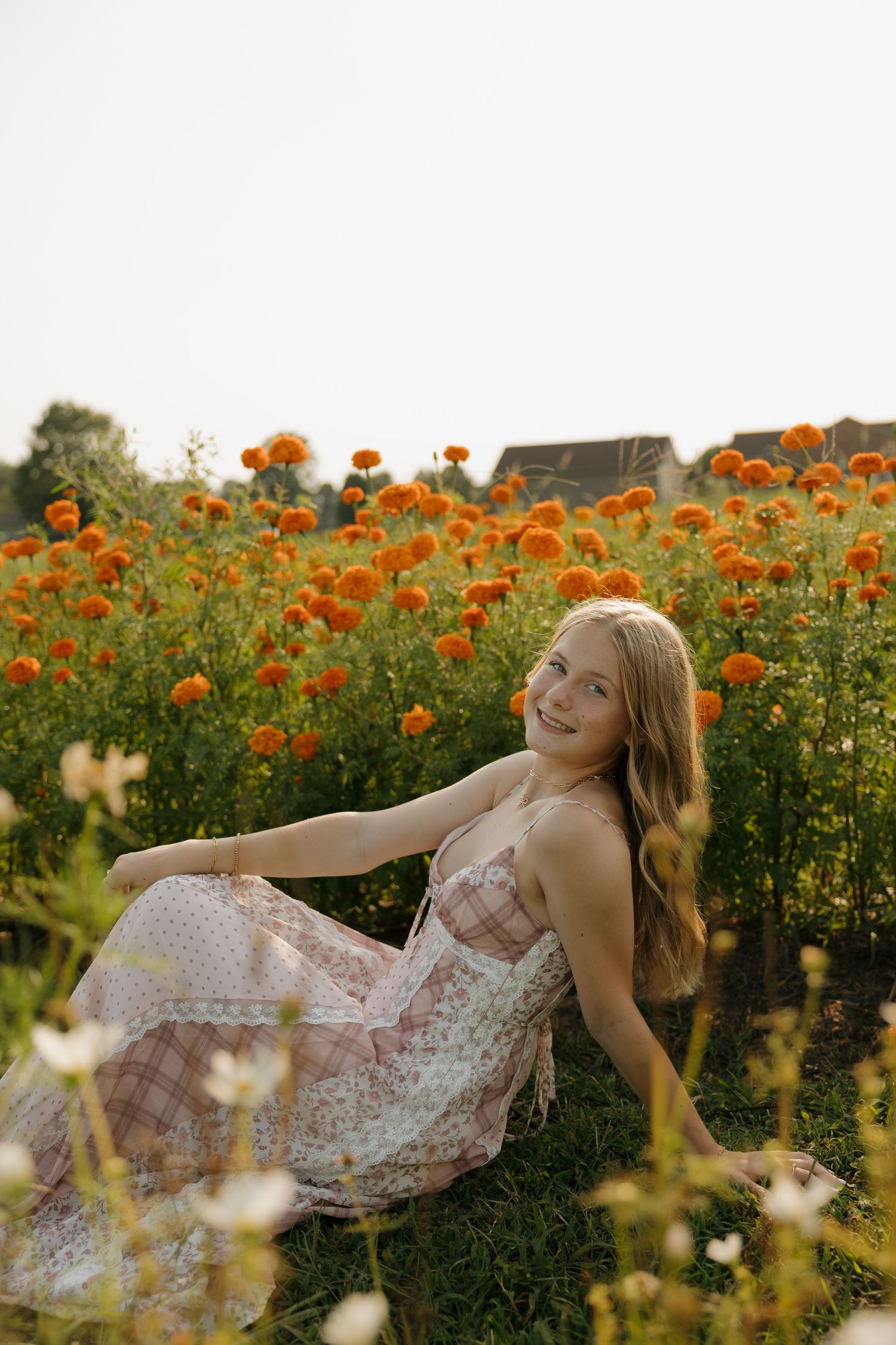 Young blonde woman in floral dress sitting in a vibrant orange marigold flower field, smiling outdoors.