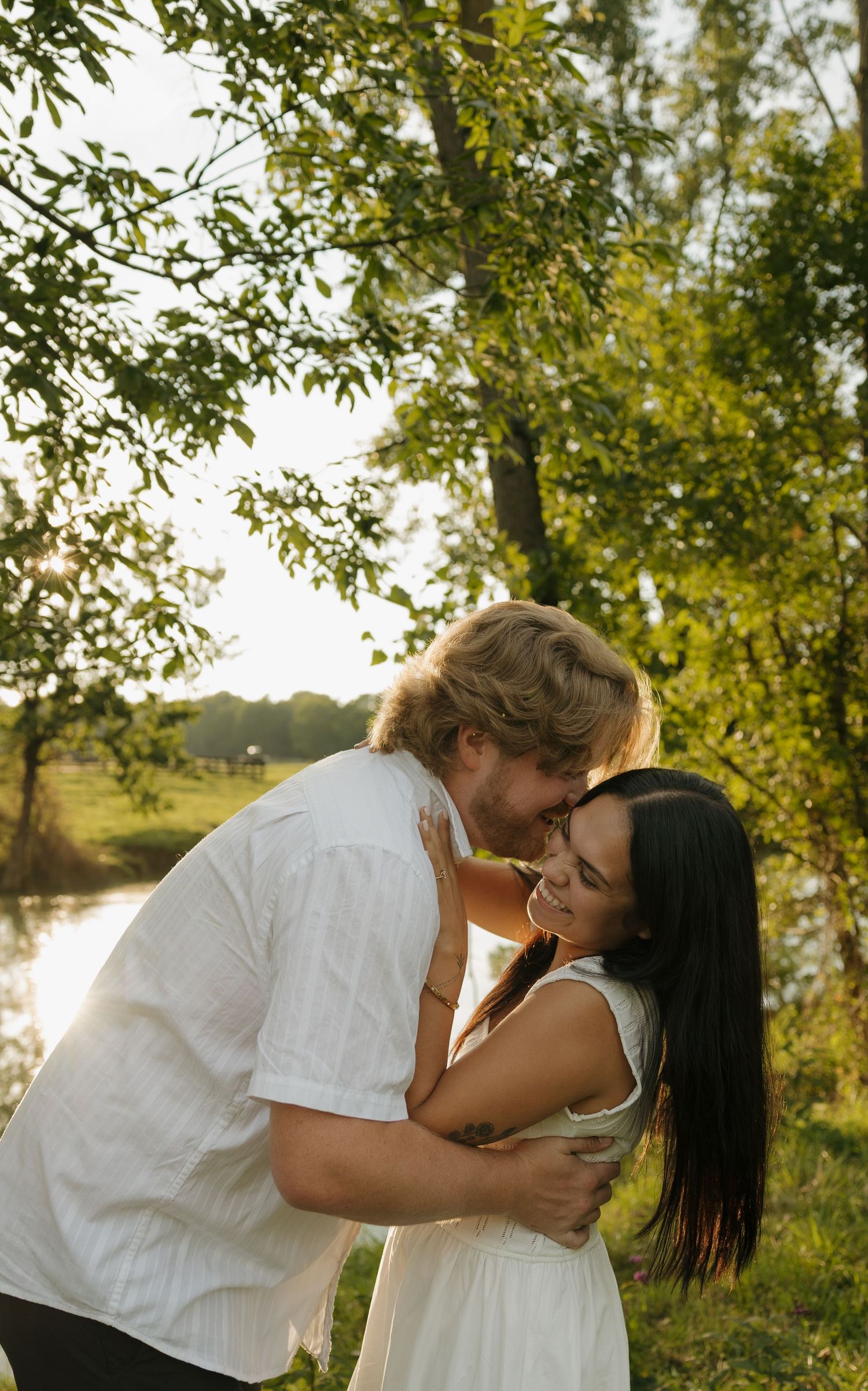 A couple embraces and shares a romantic moment outdoors near a sunlit river surrounded by lush green trees.
