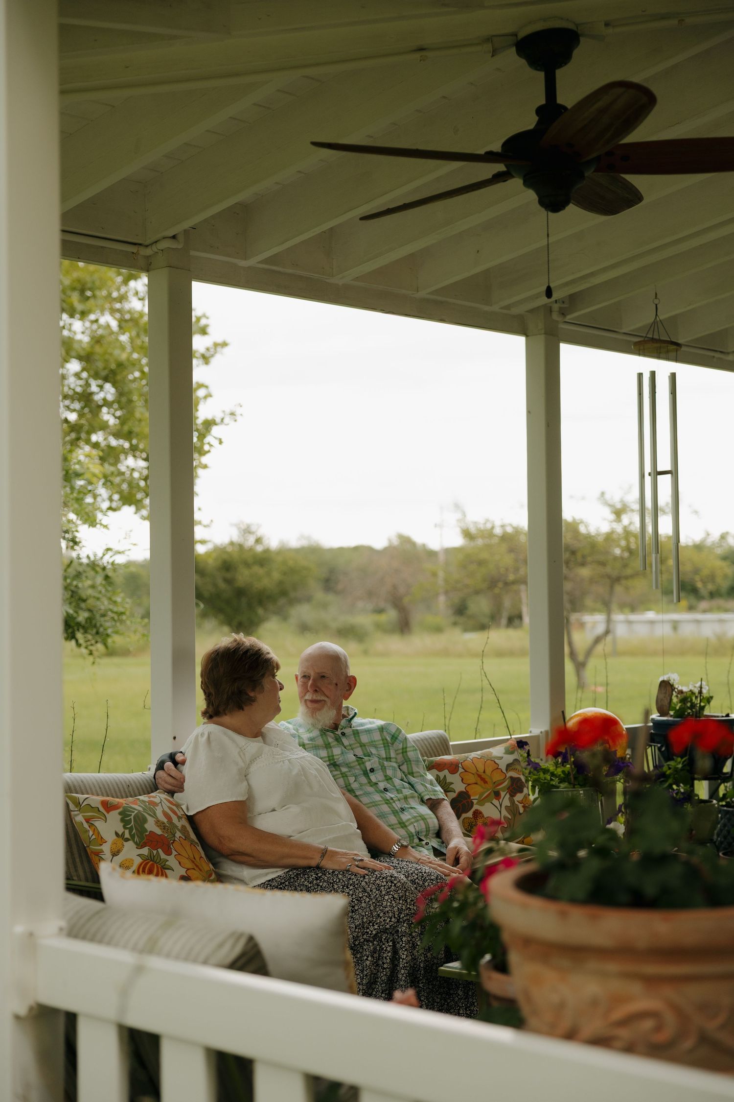 Elderly couple sitting together on a covered porch swing, enjoying a peaceful afternoon overlooking a green lawn.