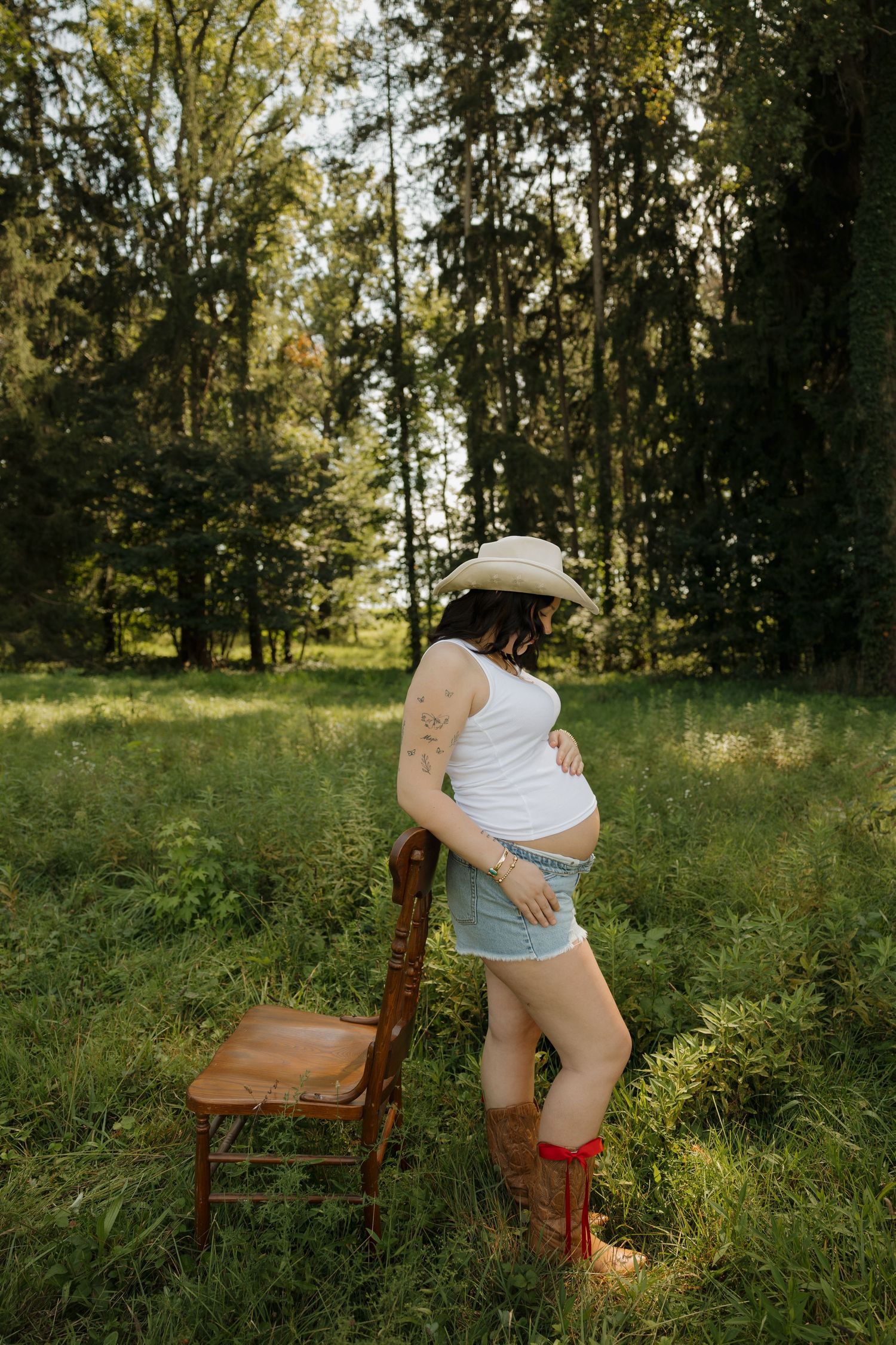 Pregnant woman in cowboy hat, white tank top, denim shorts, and red boots standing by wooden chair in green forest.