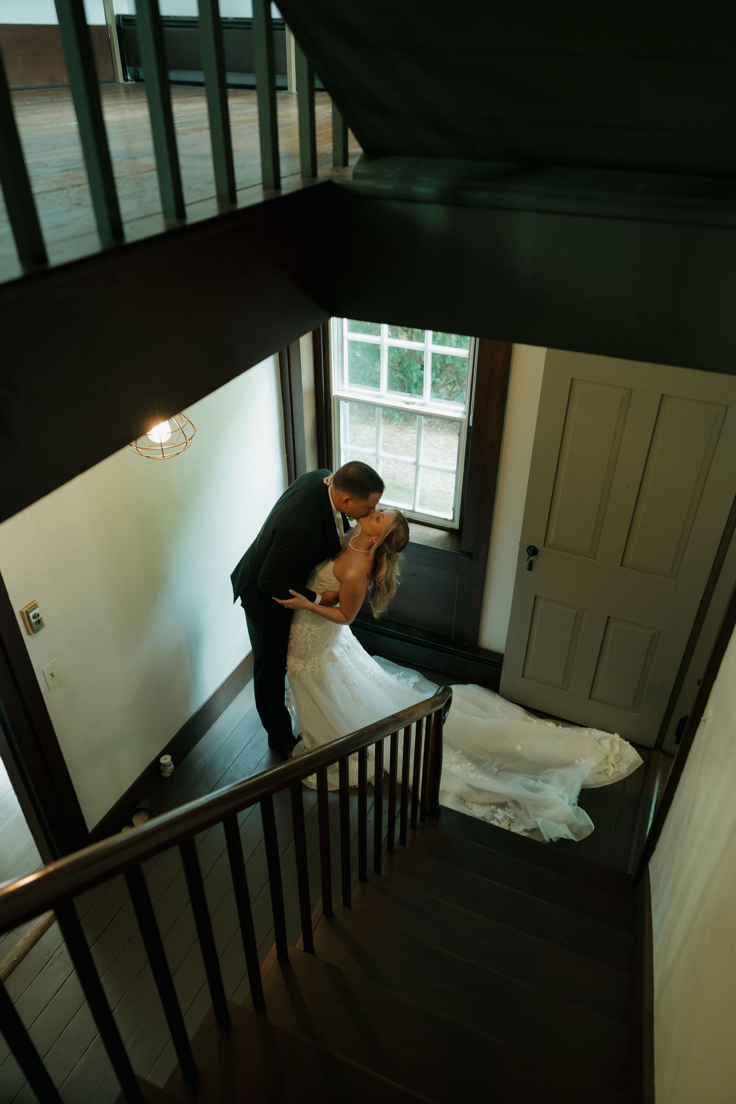 Bride and groom sharing a romantic kiss on a staircase, with wedding dress train flowing, viewed from above.