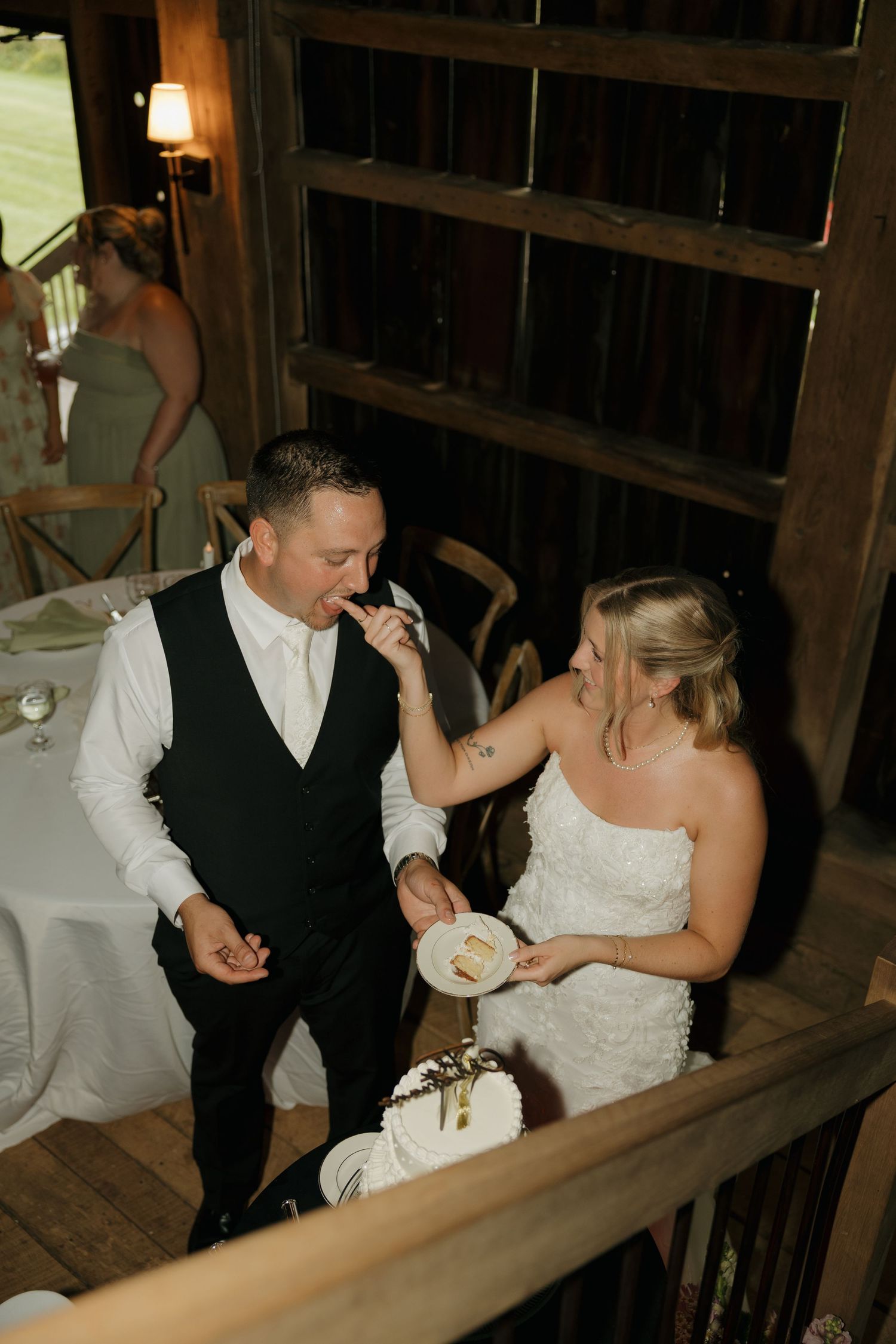Bride feeds groom wedding cake during rustic barn reception celebration indoors with warm lighting.