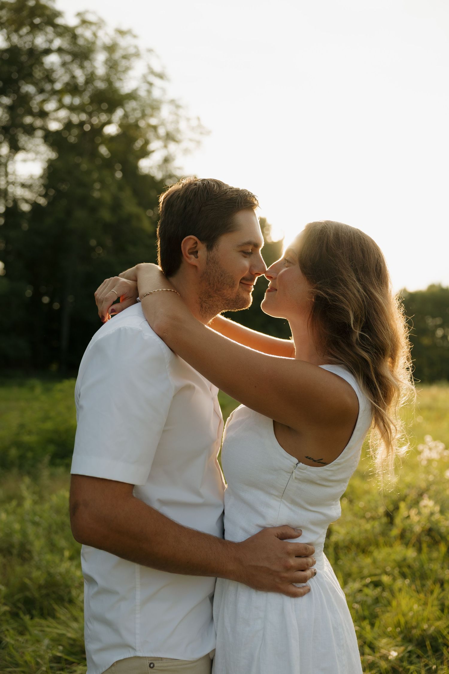 Couple sharing a romantic almost-kiss embrace in a sunlit meadow, wearing white outfits with golden hour backlight.