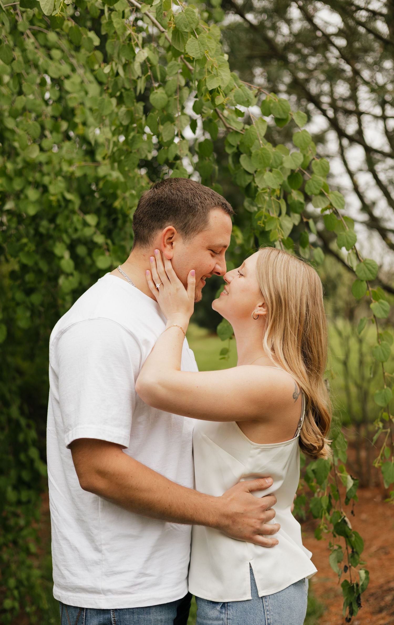 A couple shares a romantic almost-kiss embrace outdoors under lush green tree branches, wearing casual white tops.