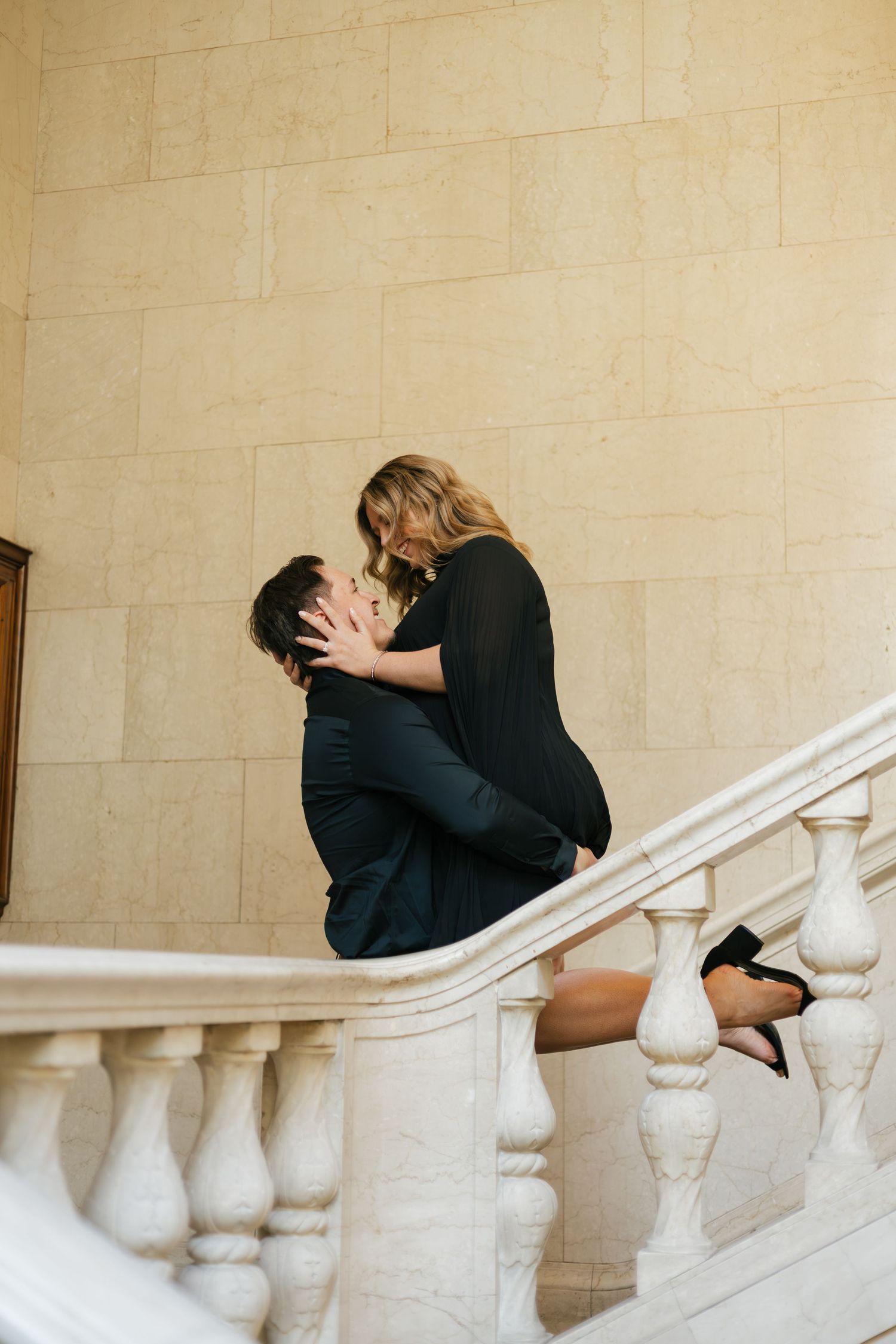 Couple sharing a romantic kiss on elegant marble staircase, man in dark suit, woman in black dress and heels.