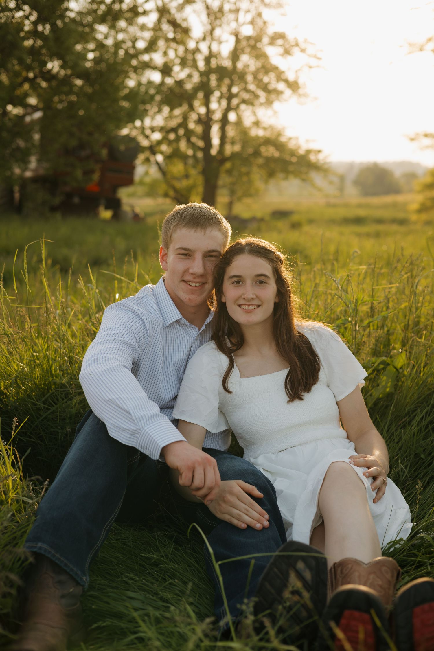 Smiling couple sitting together in lush green field at golden hour, woman in white dress and cowboy boots.