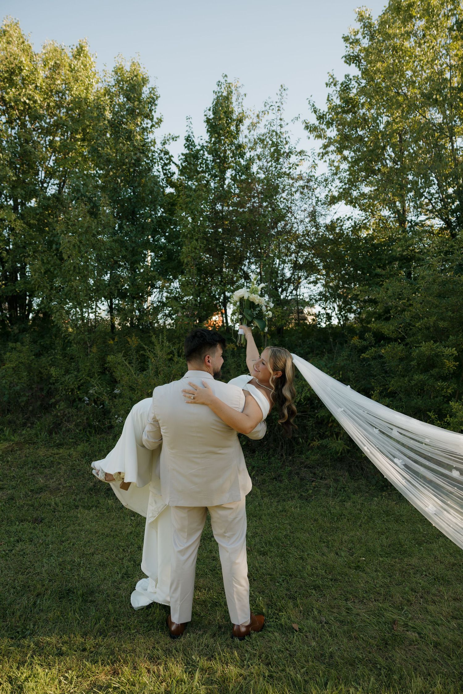 Groom carrying bride in white gown with flowing veil outdoors, surrounded by lush green trees on their wedding day.