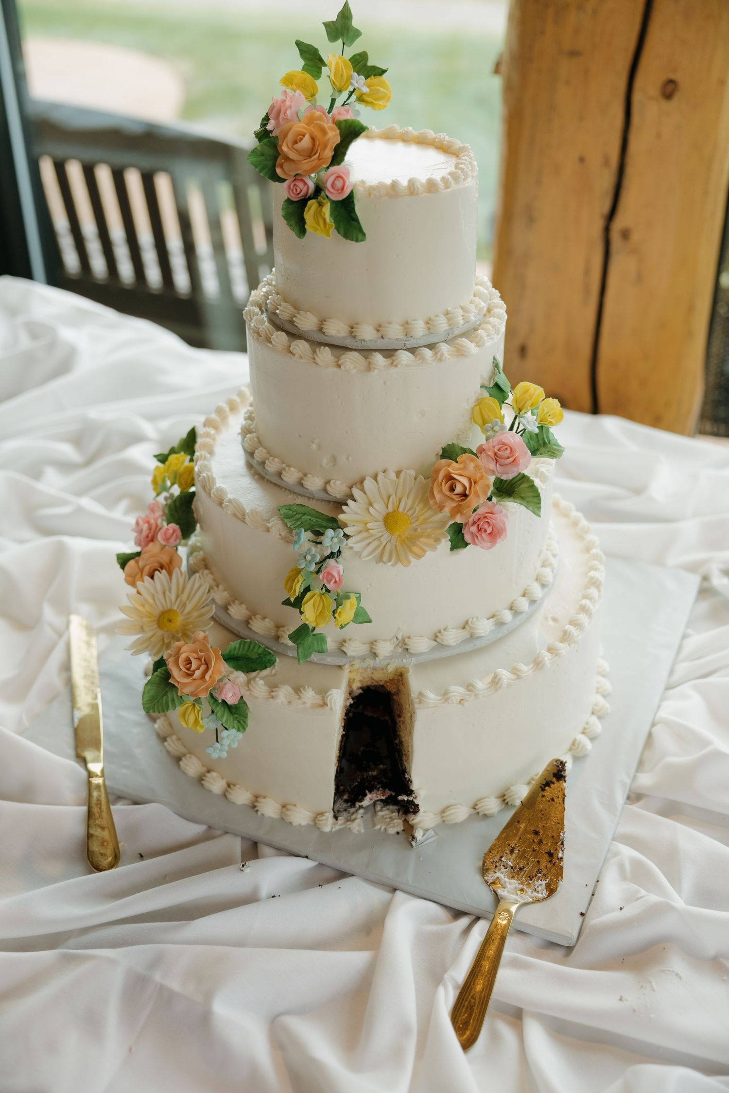 Three-tier white wedding cake with colorful sugar flowers, pearl borders, and chocolate interior revealed by a cut slice on draped table.