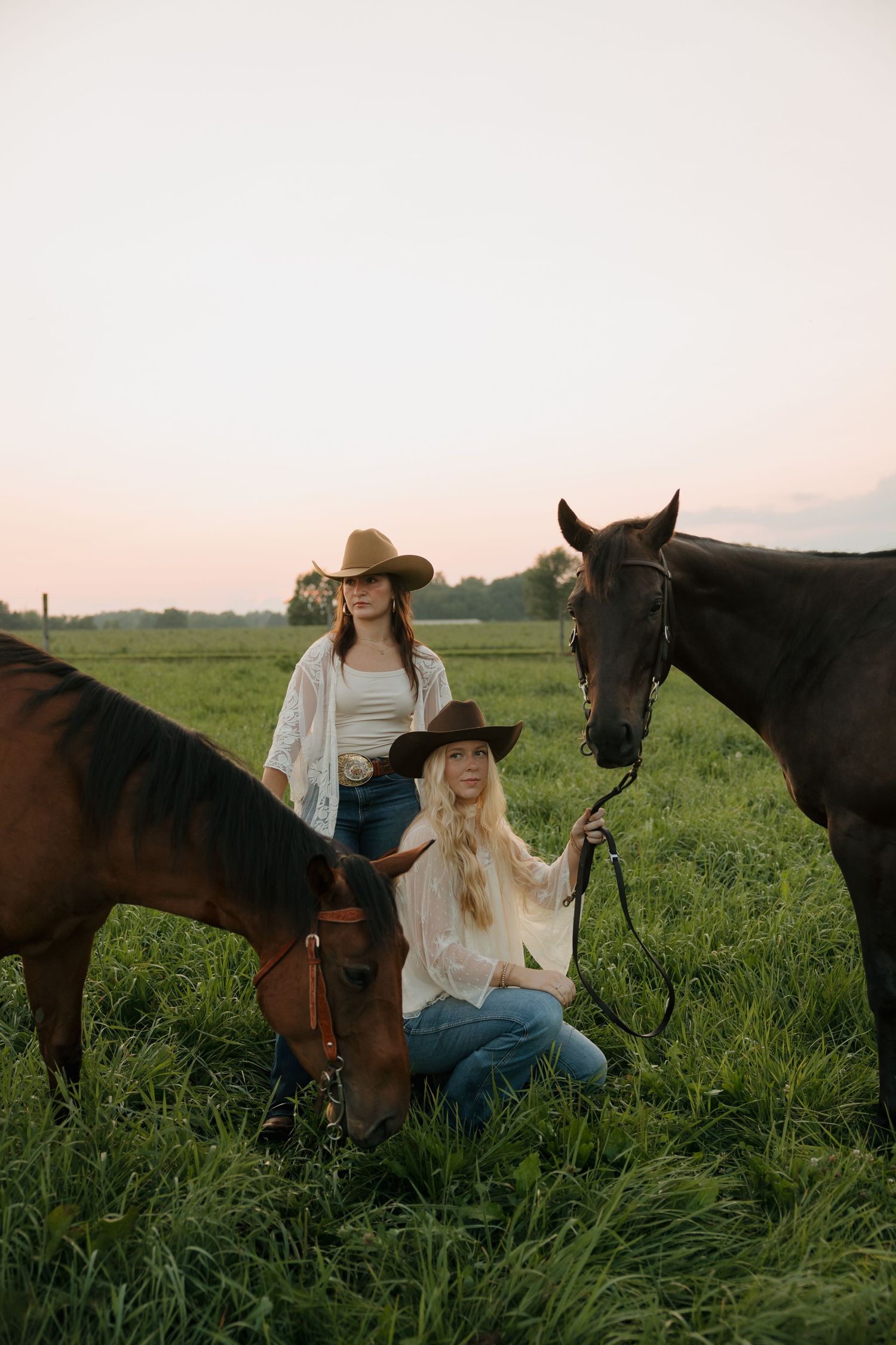 Two cowgirls in hats sit with brown horses in a lush green field at sunset.