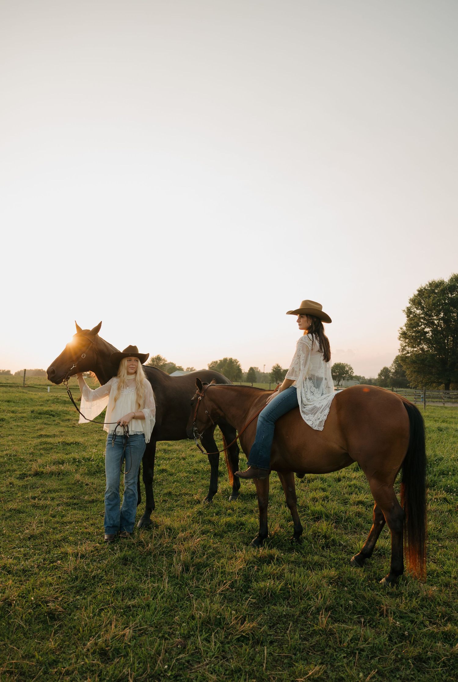 Two women in cowboy hats and white tops with jeans pose with brown horses in a golden sunset field.
