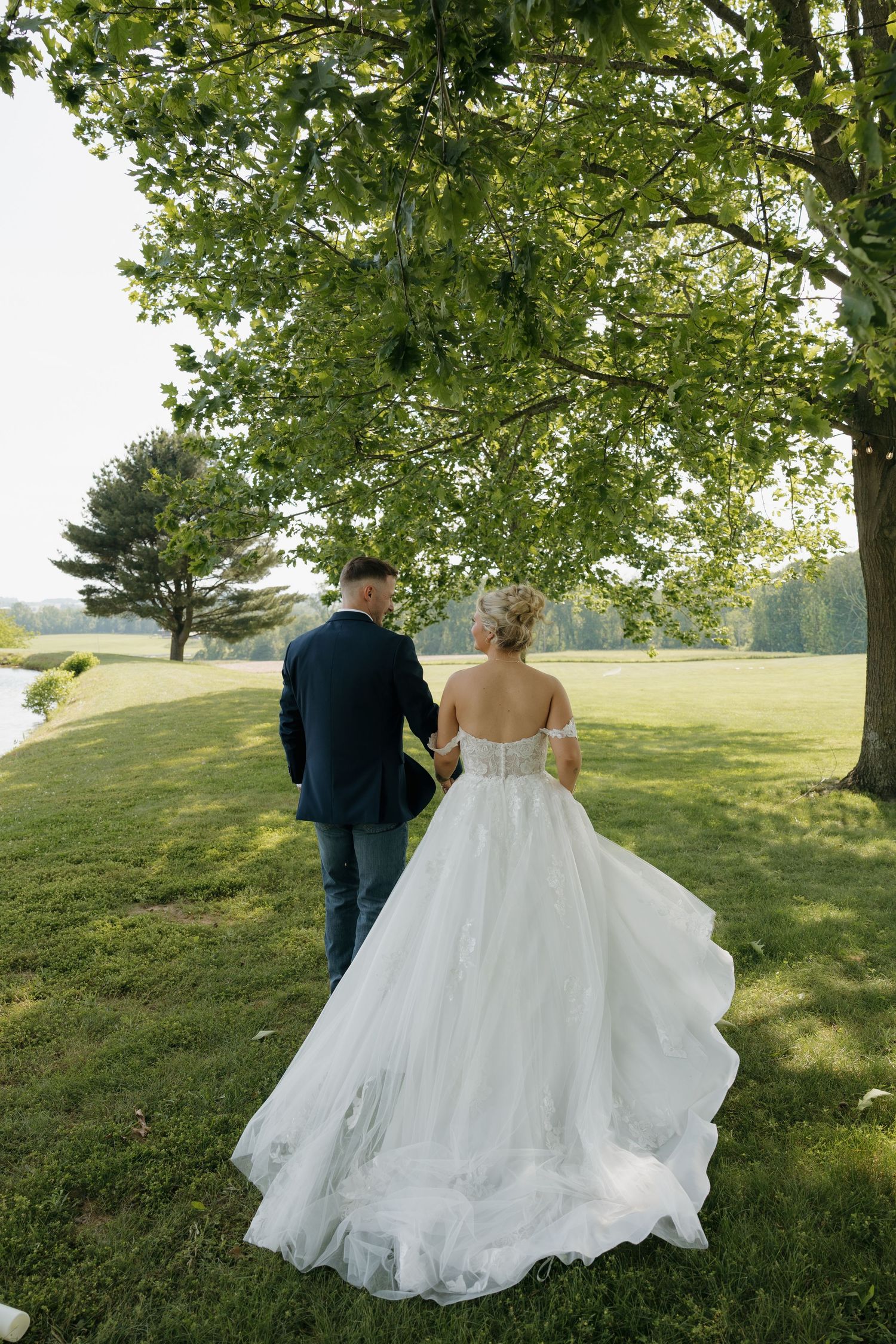 Bride and groom walking hand in hand under a large green tree on a sunny day, bride wearing a flowing white ballgown.