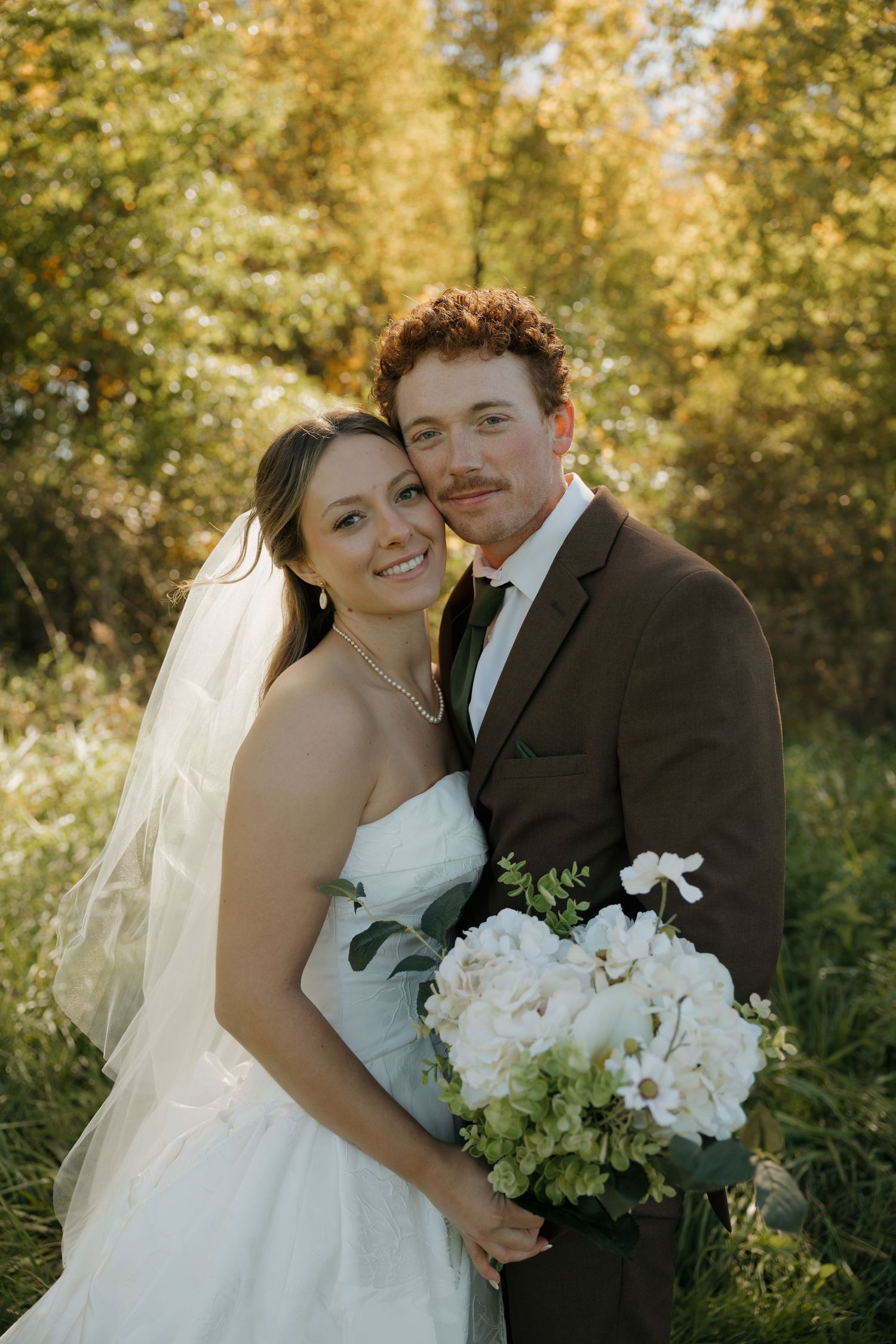 Smiling bride in a strapless white gown and veil holds a white hydrangea bouquet beside her groom in a brown suit among autumn trees.