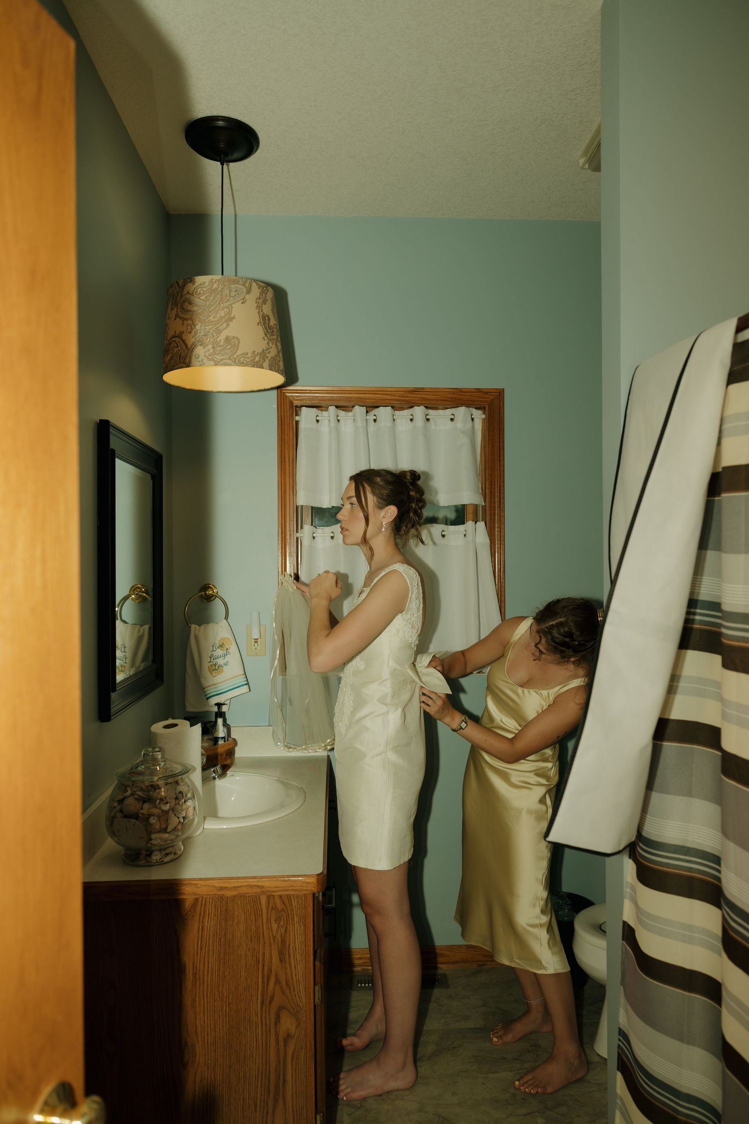 Two women in a bathroom, one in a white dress being helped by another in a gold dress near a sink.