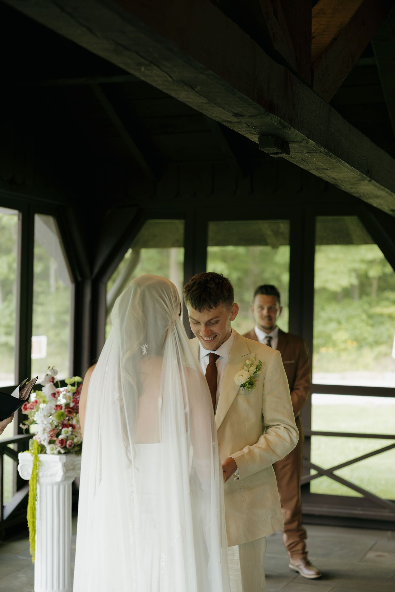 Groom in beige suit smiling at bride in white veil during outdoor wedding ceremony in rustic pavilion.