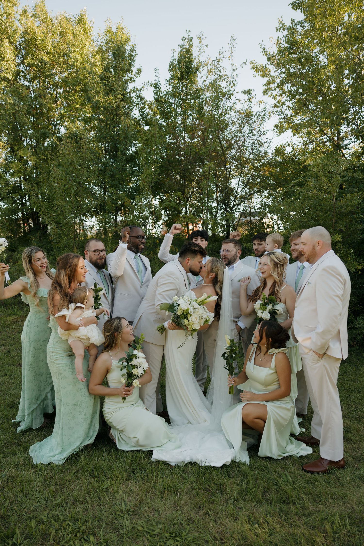 Wedding party celebrates outdoors with bride and groom kissing, bridesmaids in sage green gowns and groomsmen in tan suits.