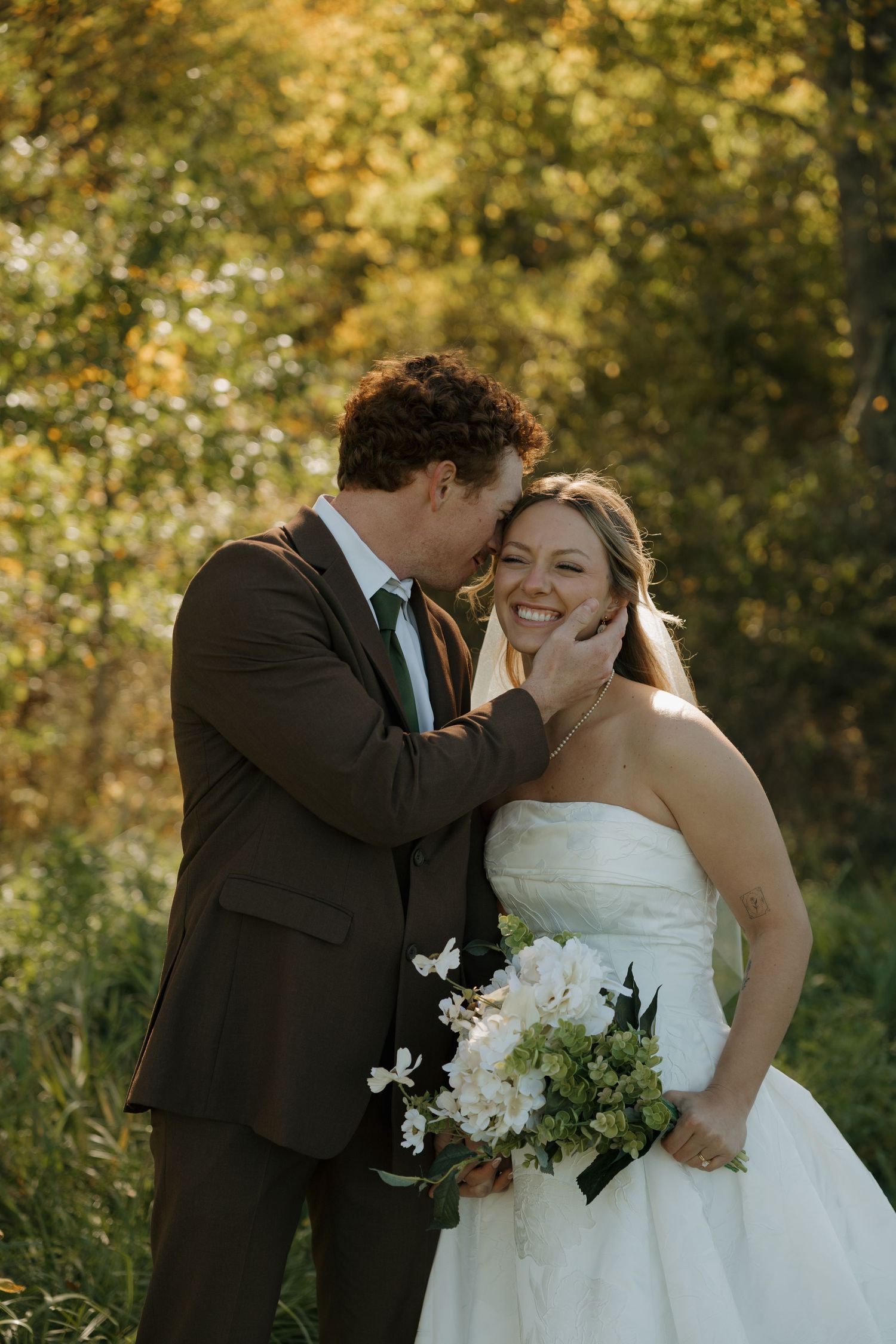 Smiling bride in a strapless white gown holding a white floral bouquet as her groom in a brown suit kisses her cheek outdoors in autumn.