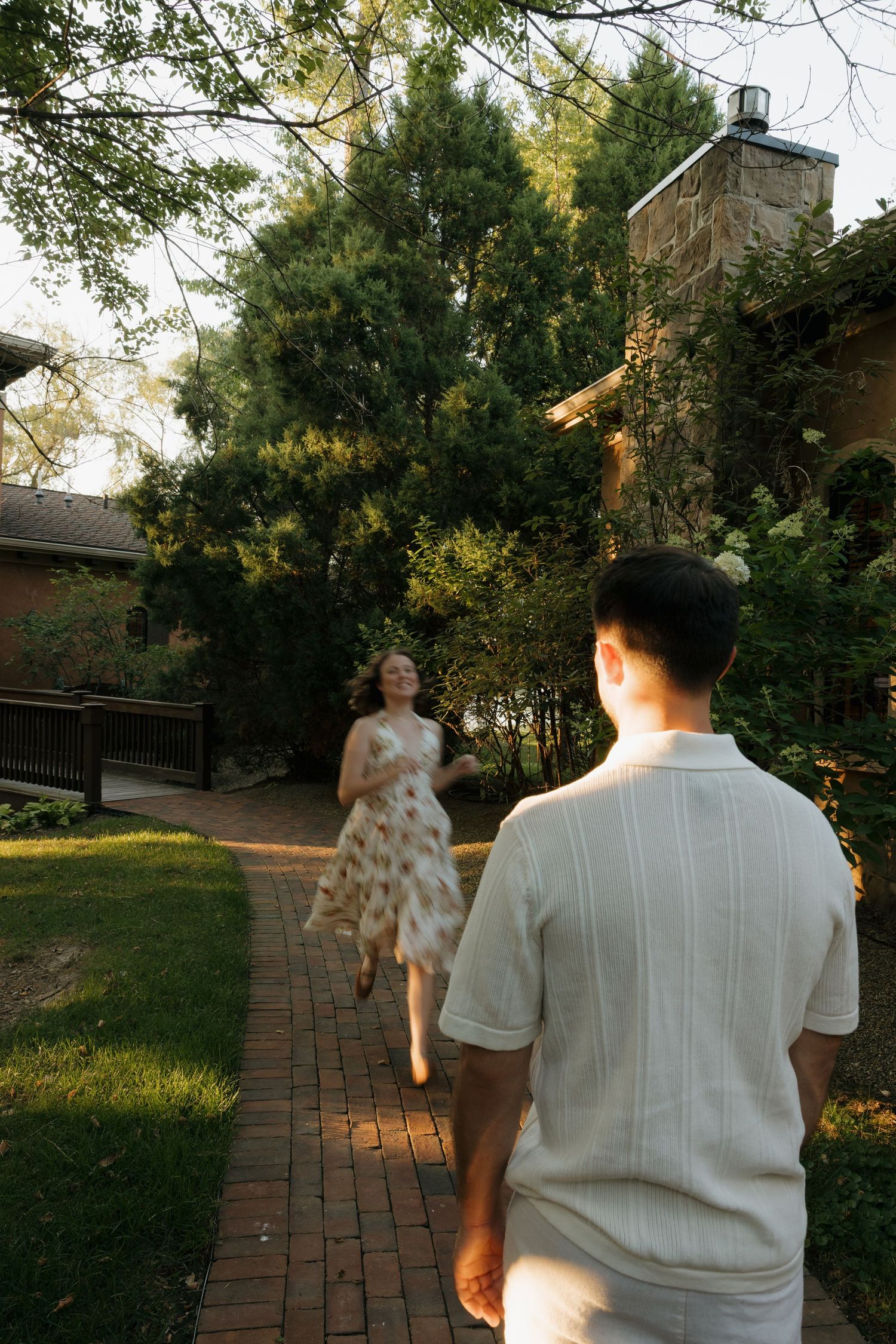 A woman in a floral dress runs toward a man on a brick garden path surrounded by lush green trees.