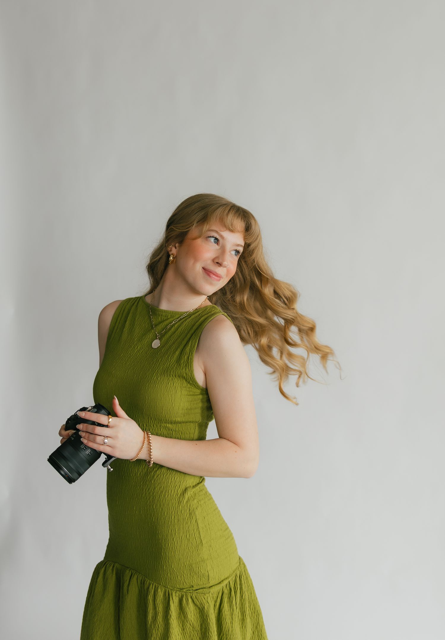 Young woman in olive green dress holding a camera, with flowing blonde hair against a white studio background.