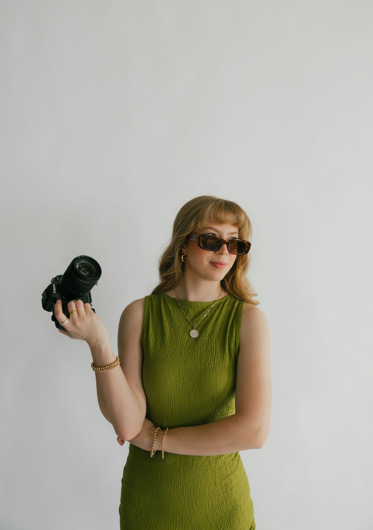 Woman in green dress holding camera with sunglasses on white background.