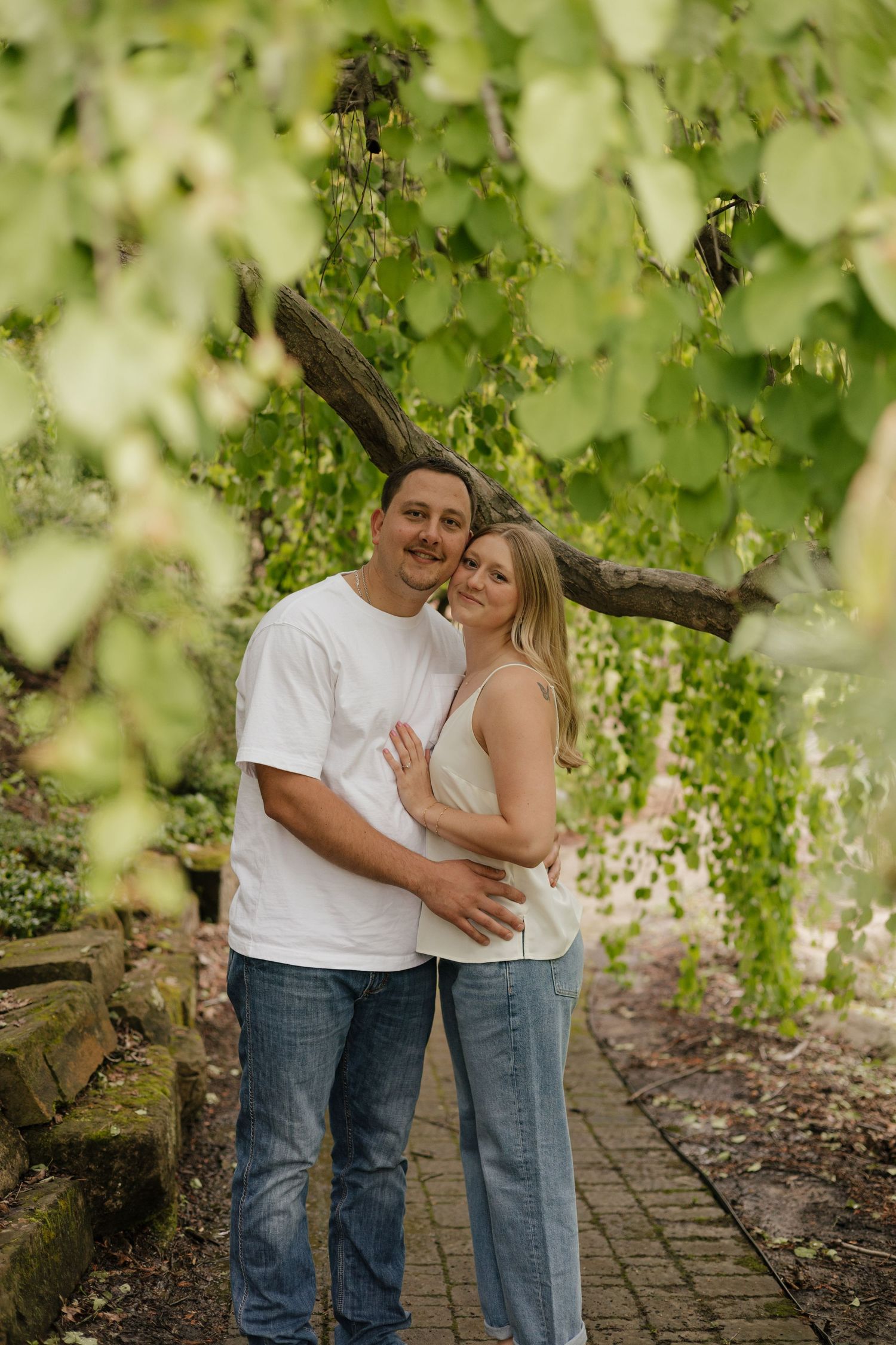 Couple embracing and smiling outdoors under lush green tree branches on a garden stone path.