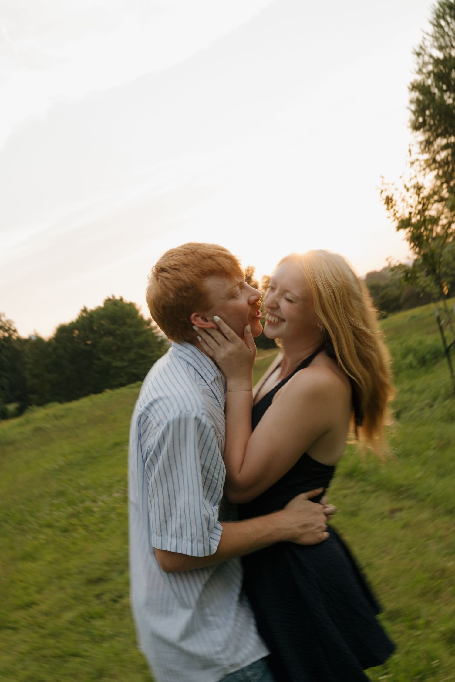 Couple laughing and kissing outdoors at sunset in a green meadow, woman in black dress and man in striped shirt embracing.