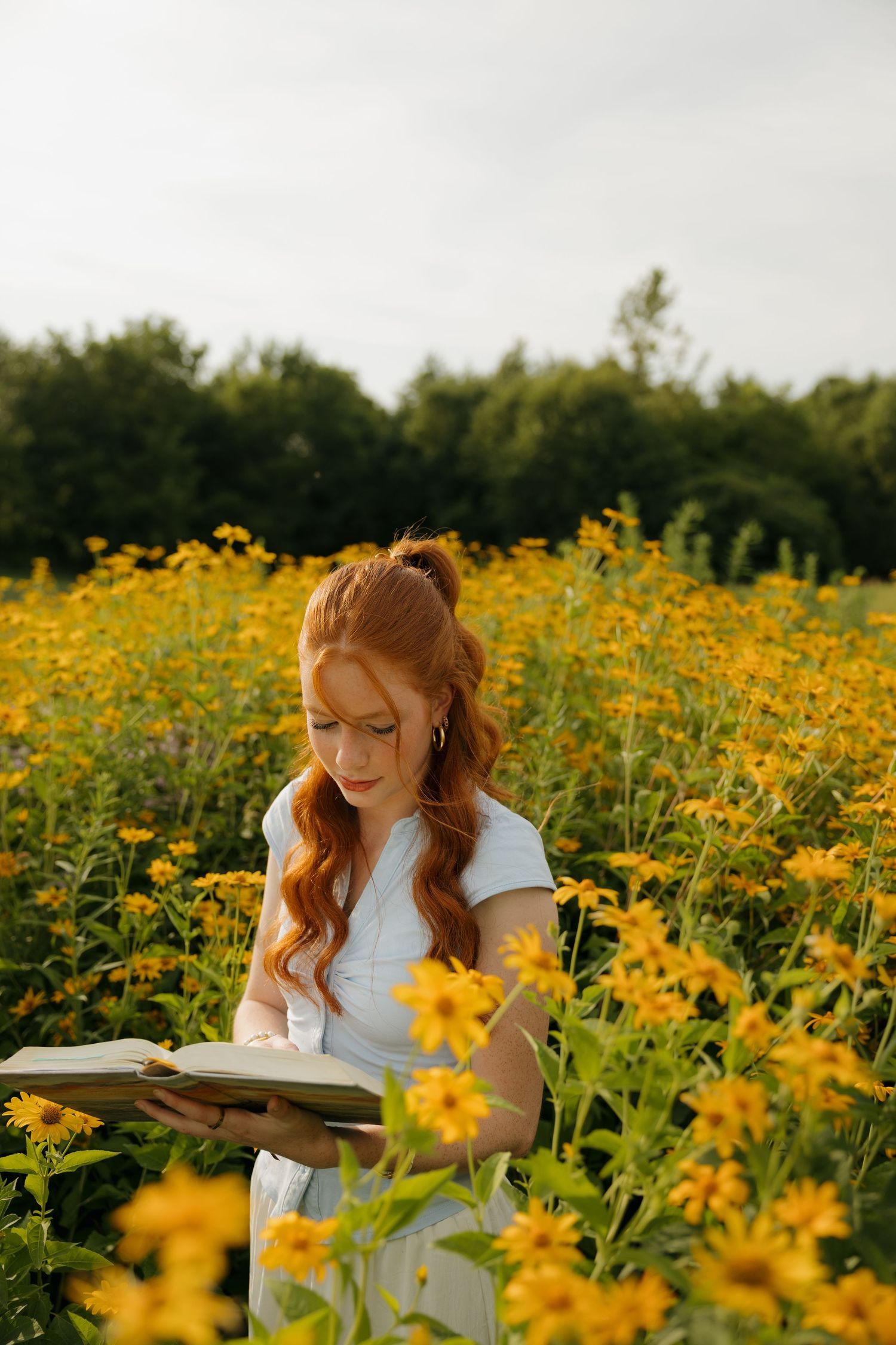 Red-haired woman in white dress reading a book in a vibrant yellow wildflower field outdoors.