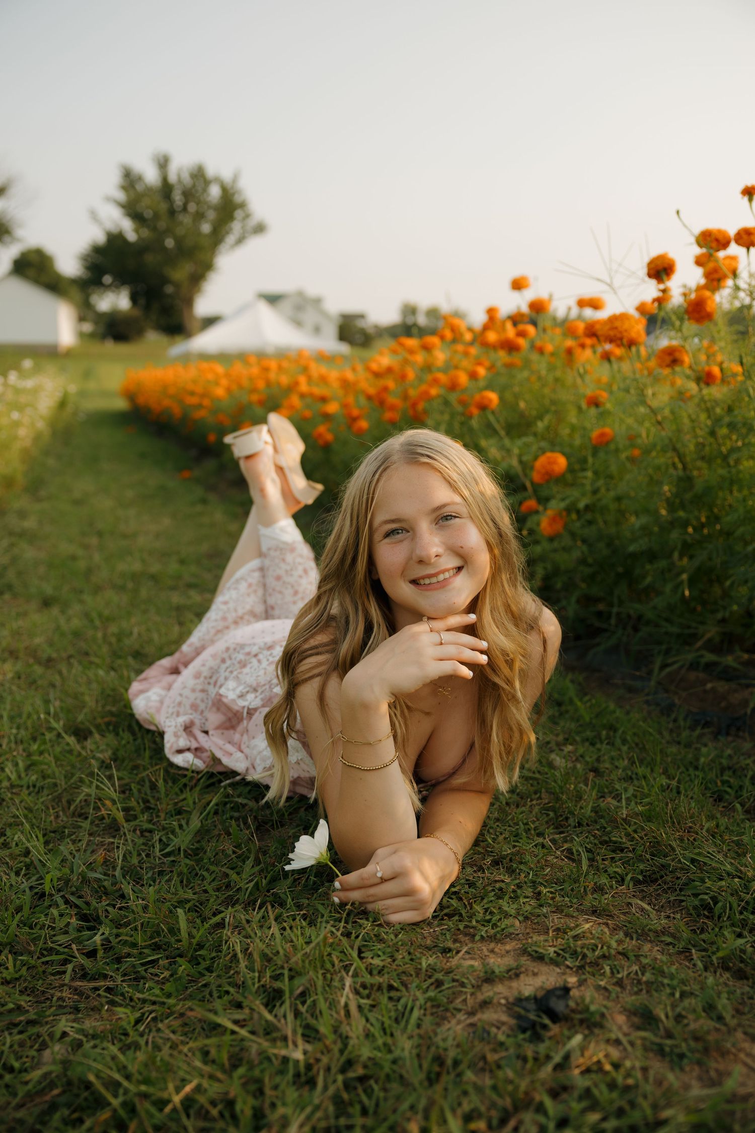 Young blonde girl lying on grass smiling, surrounded by vibrant orange marigold flowers in a sunny field.