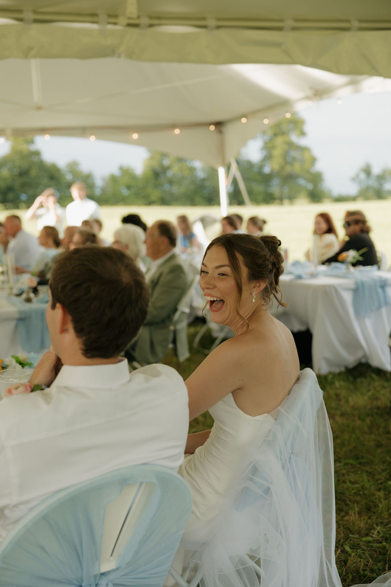 Bride laughing joyfully at outdoor wedding reception under white tent with string lights and guests.