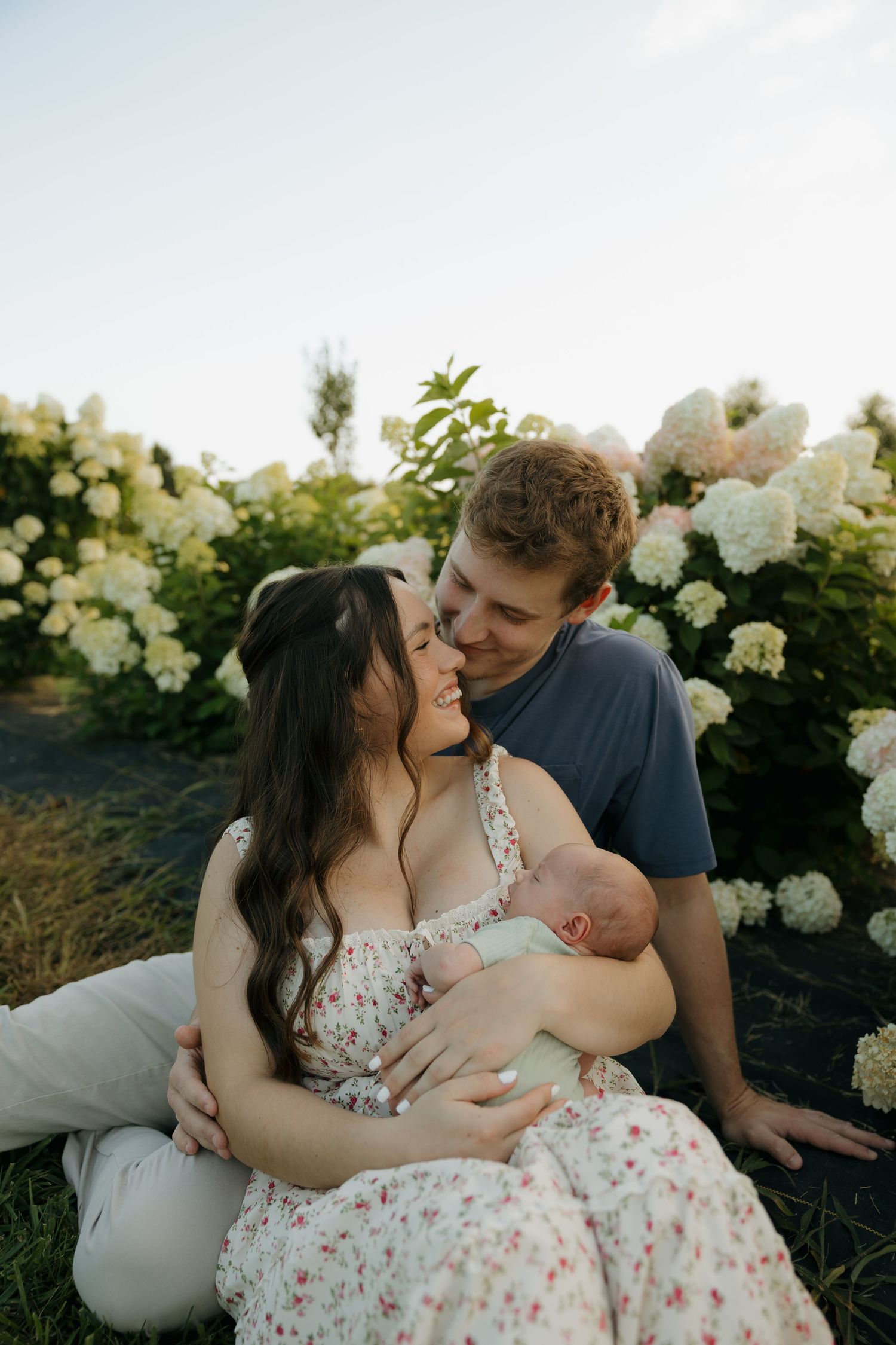 Young couple sitting in hydrangea garden, smiling and holding newborn baby in floral dress outdoors.