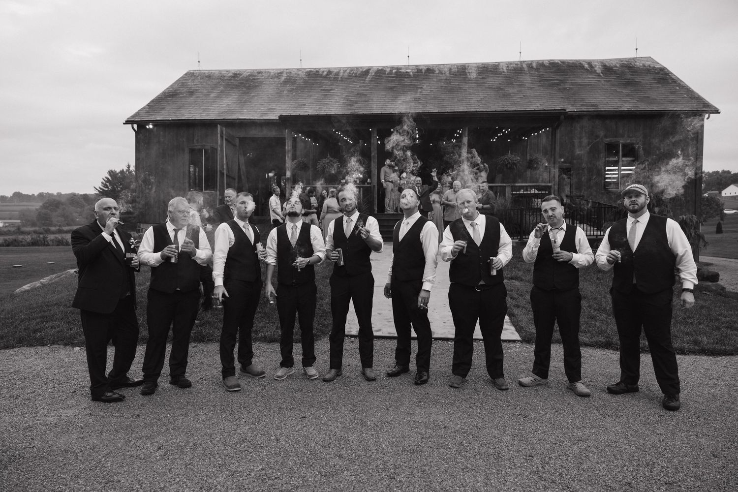 Black and white photo of groomsmen in vests and ties posing in front of a rustic barn at a wedding.