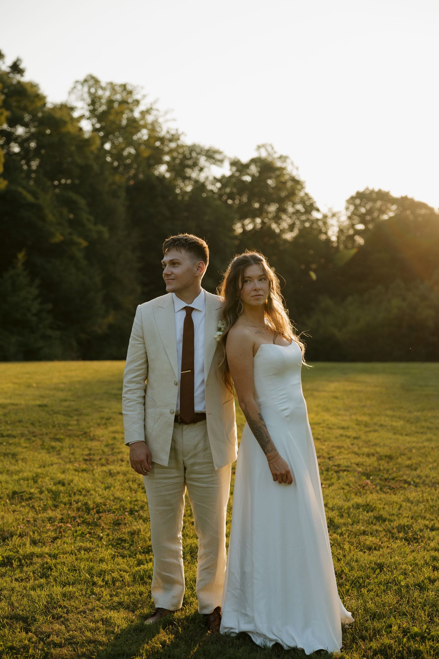 Bride and groom standing in a sunlit field at golden hour, surrounded by lush green trees at their outdoor wedding.