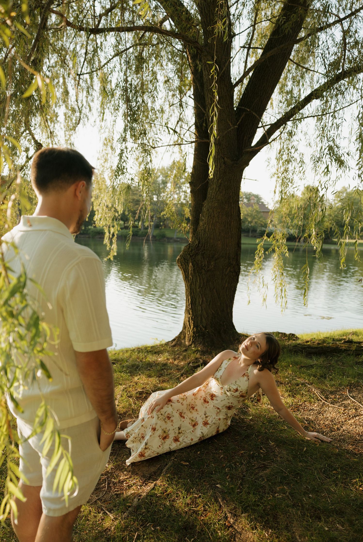 Couple enjoying a sunny day by a willow tree near a lake, woman in floral dress reclining on grass.