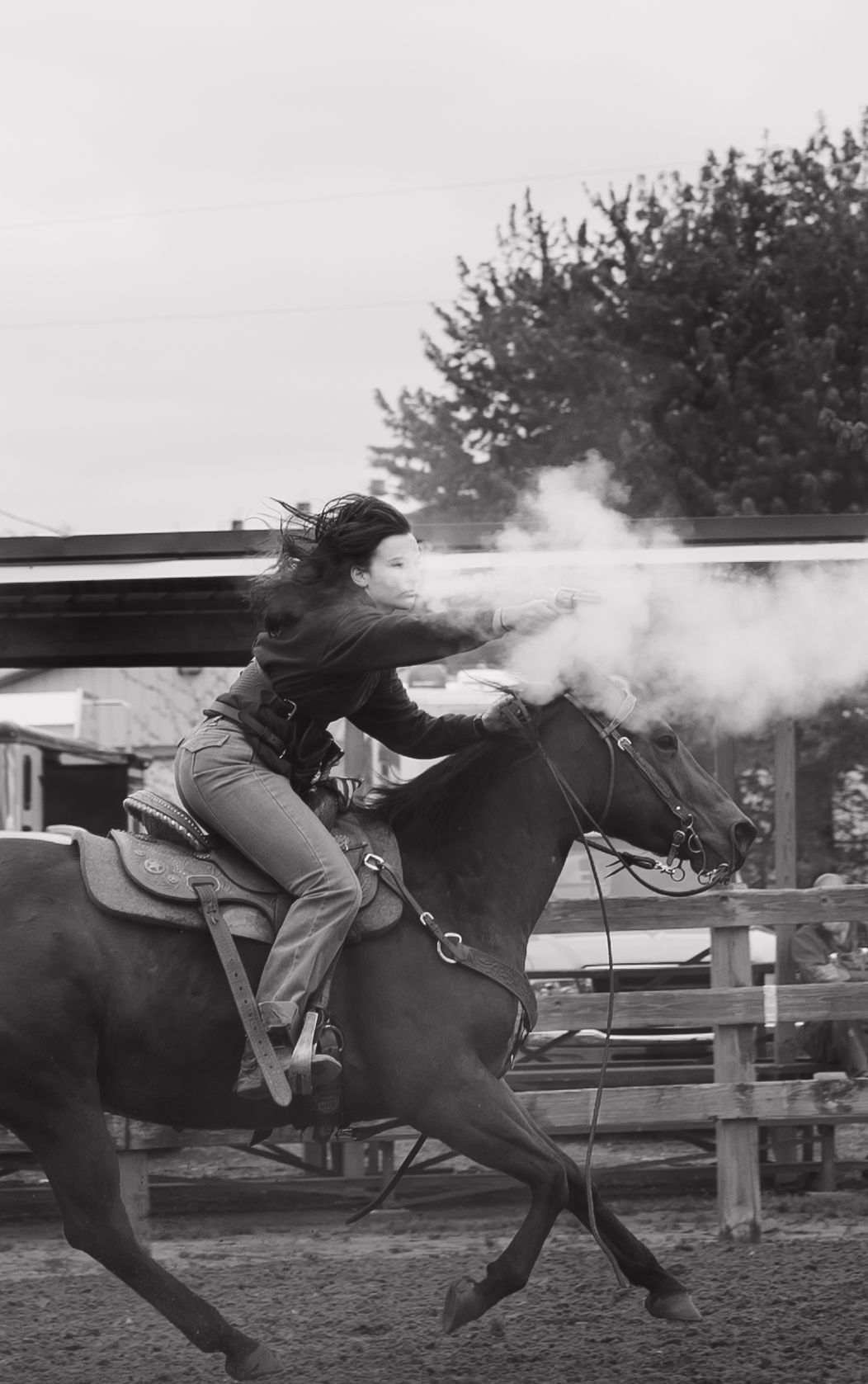 Woman riding horse firing gun in black and white rodeo action shot with smoke visible.