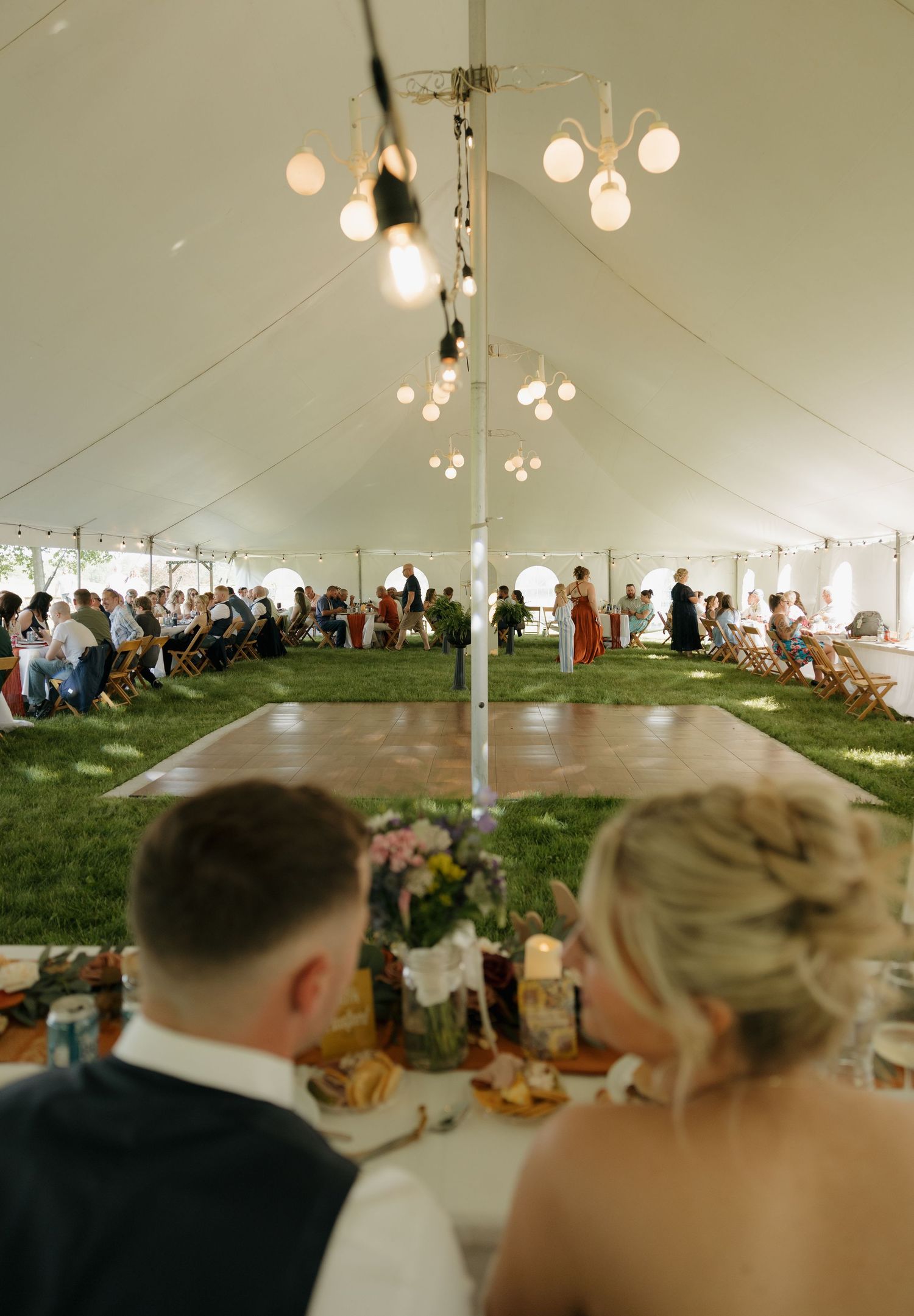 Bride and groom seated at head table overlooking elegant outdoor tent wedding reception with dance floor and string lights.