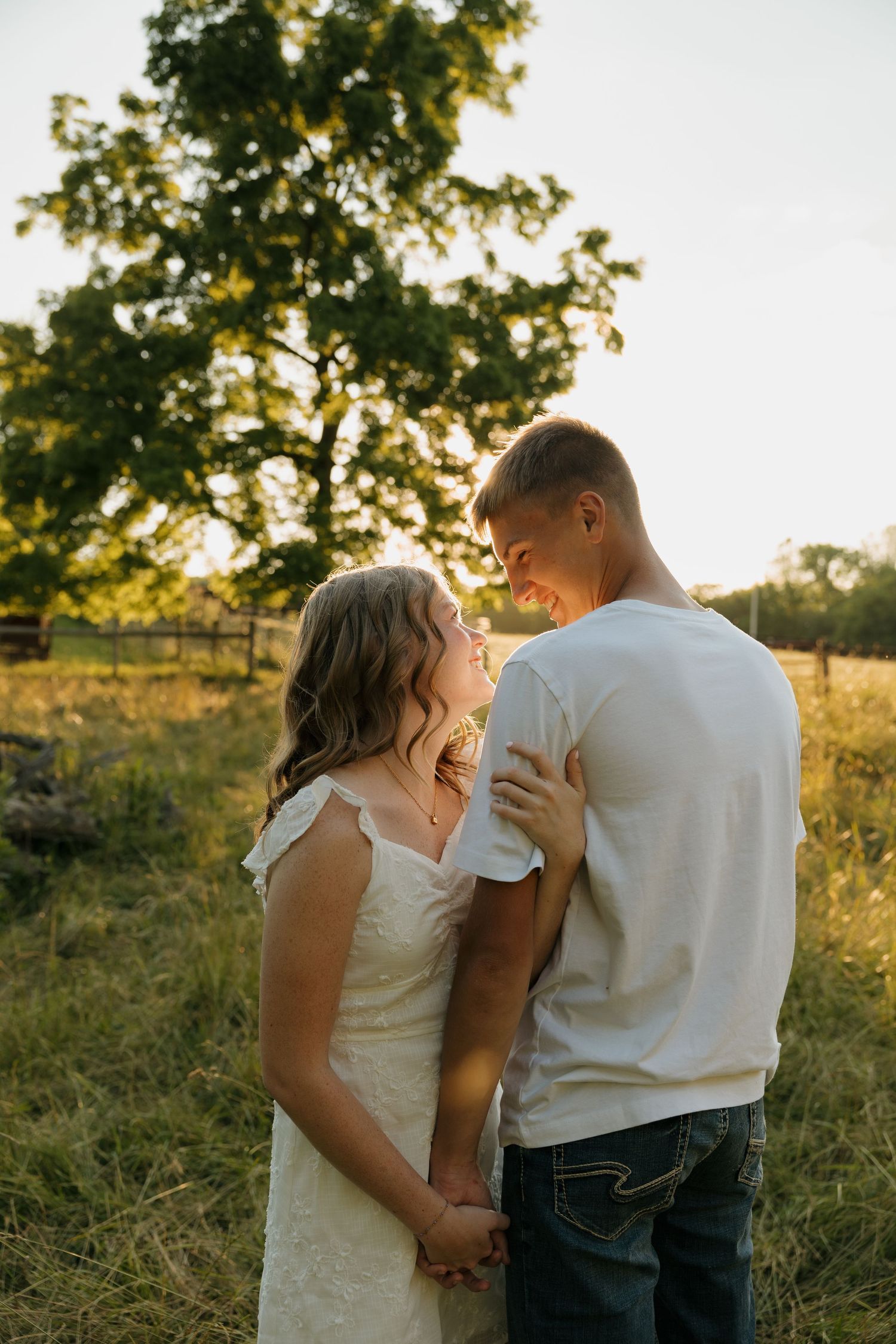 Couple in white dress and jeans holding hands and smiling at each other in a sunlit green field.