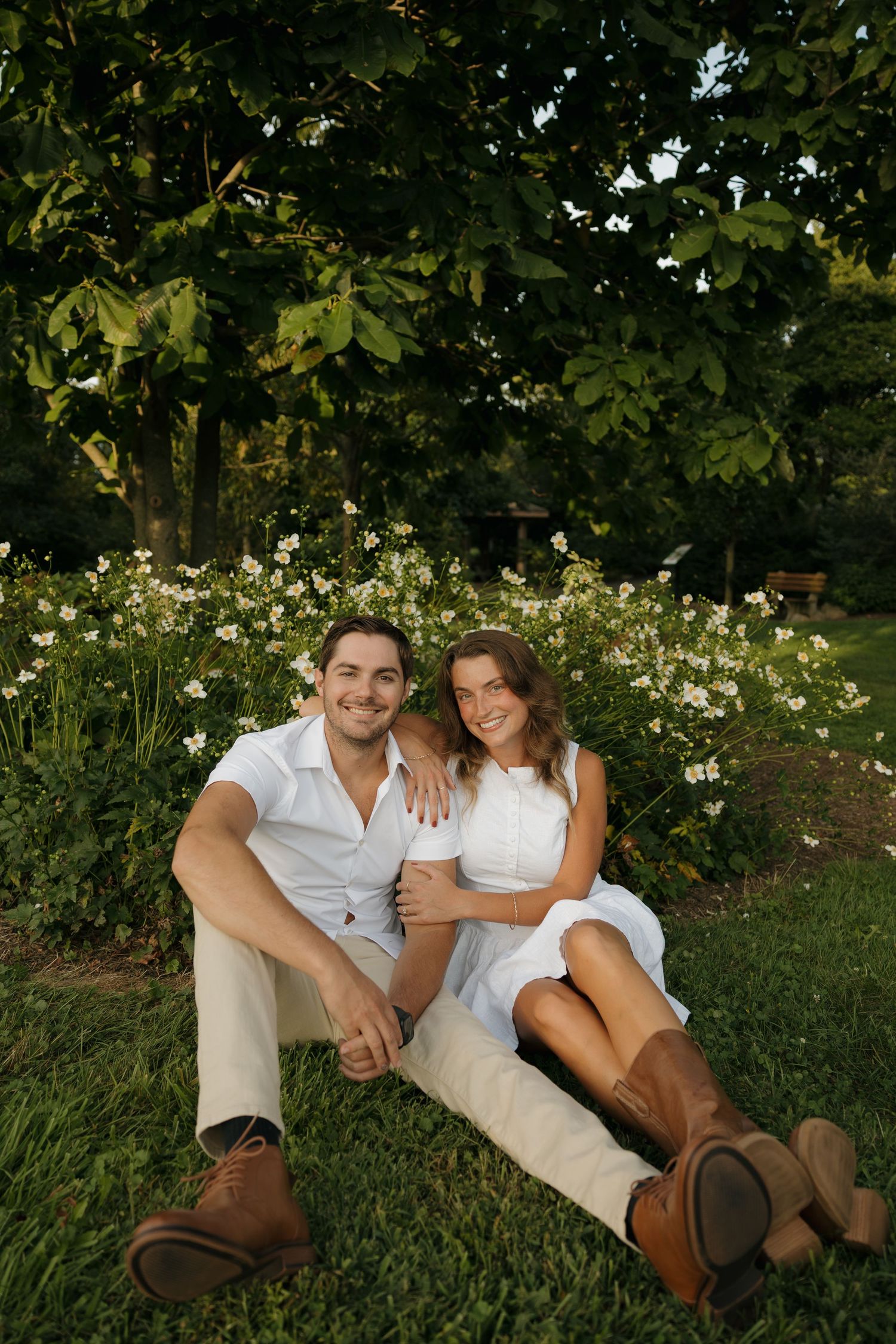 A smiling couple in white outfits sits on green grass in front of blooming white flowers and lush trees.