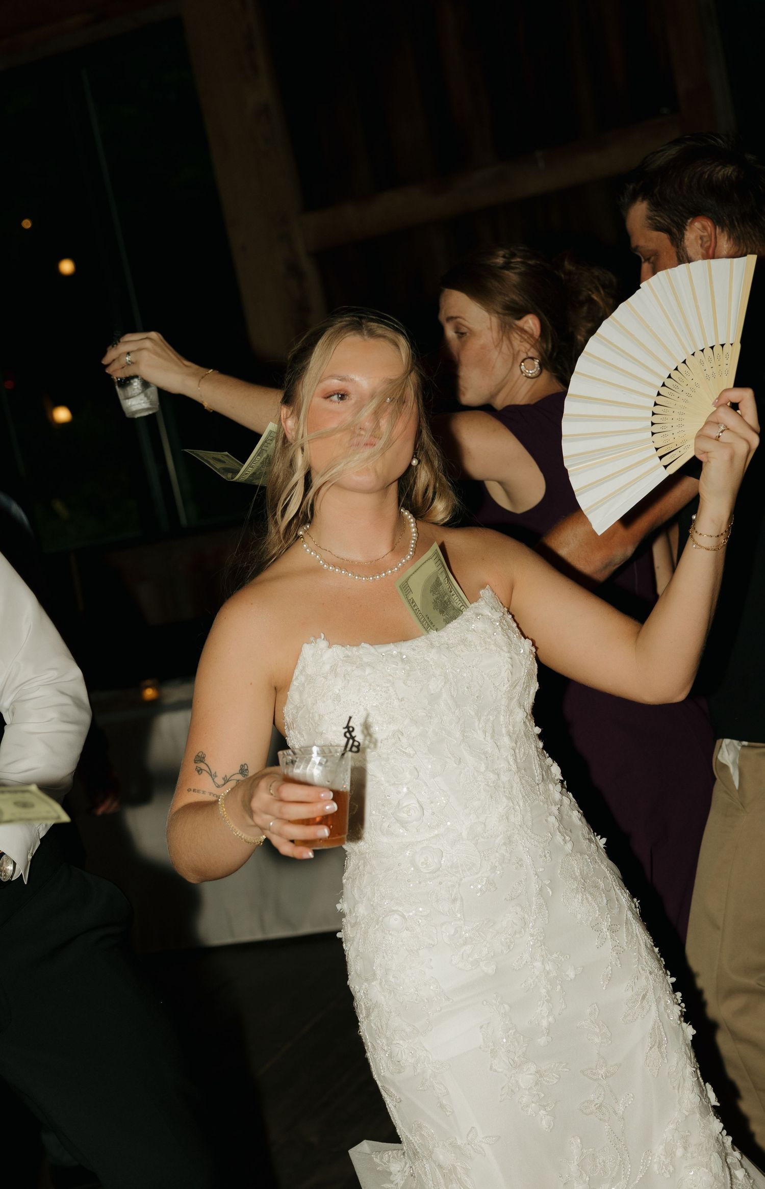 Bride in white lace gown dancing at wedding reception, holding beer, with money tucked in dress and hand fan nearby.
